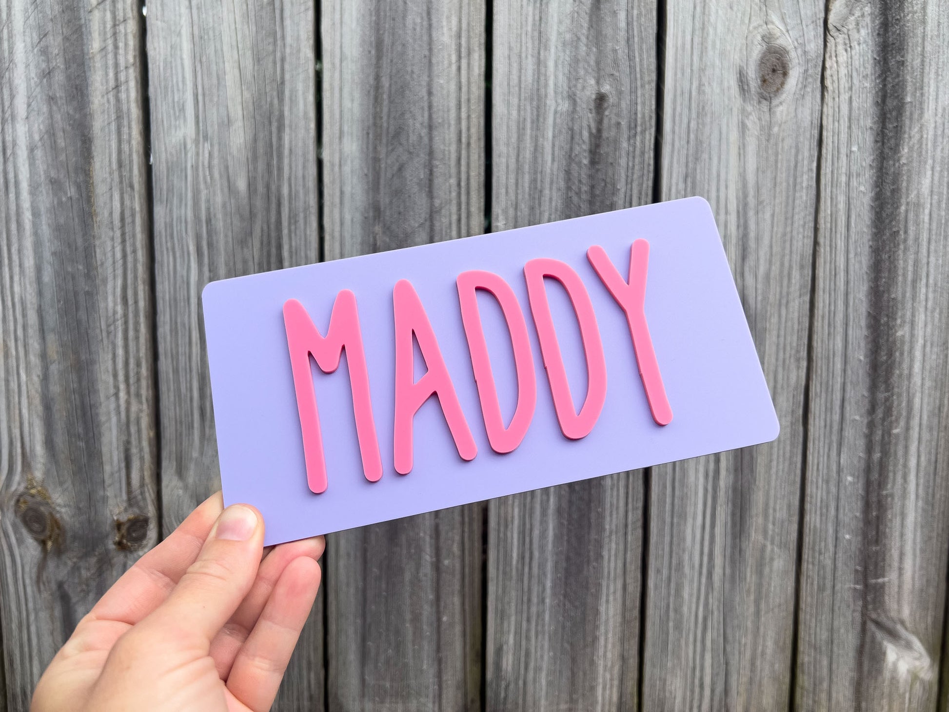 Hand holding a purple sign with 'MADDY' in pink letters against a wooden background | Coji Designs