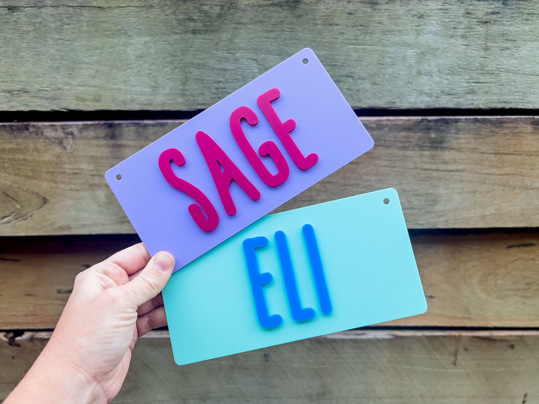 Two nameplates, one purple with 'SAGE' and one blue with 'ELI', held against a wooden background | Coji Designs