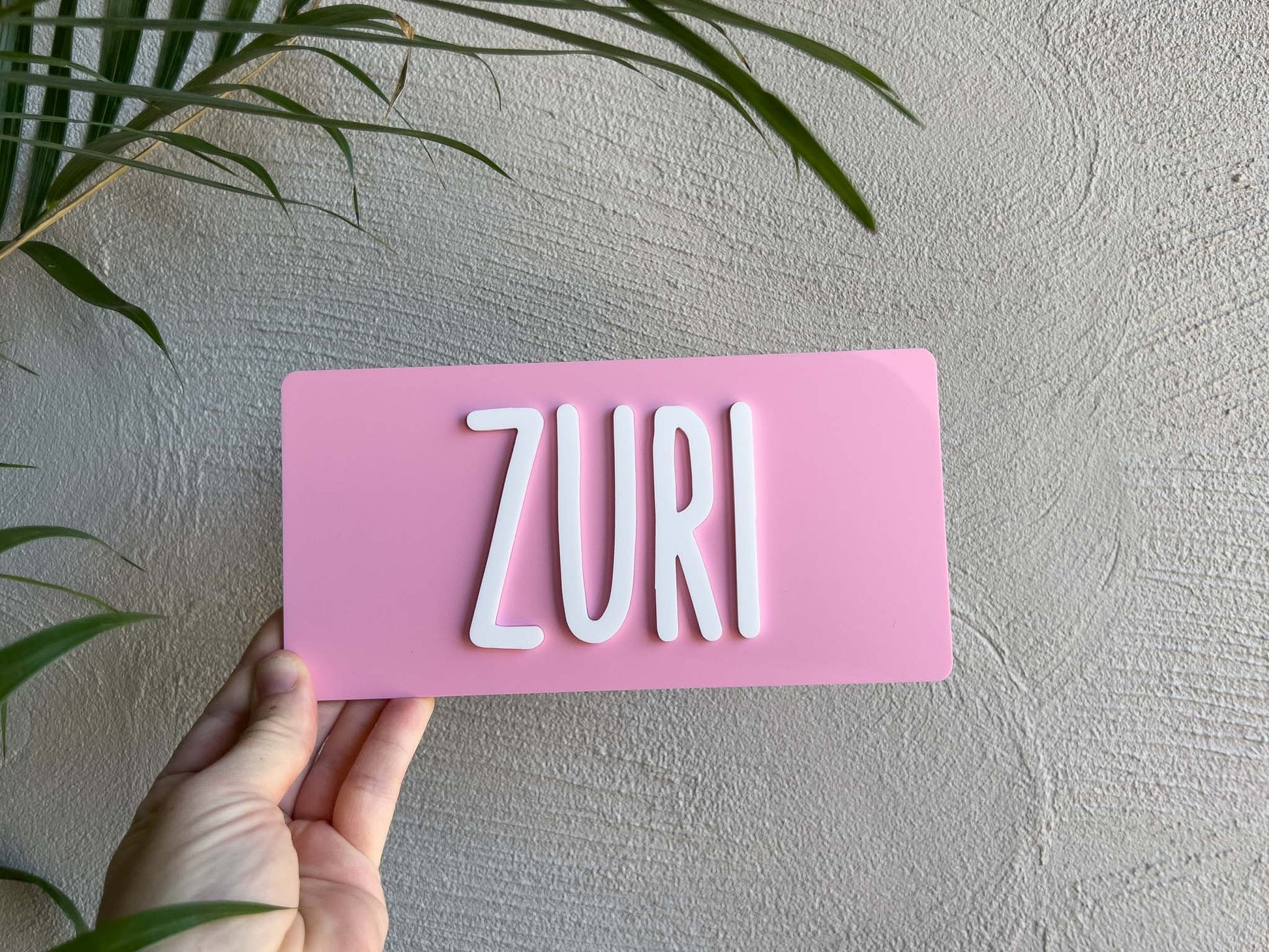 Pink rectangular sign with 'ZURI' on a light gray wall with green leaves | Coji Designs