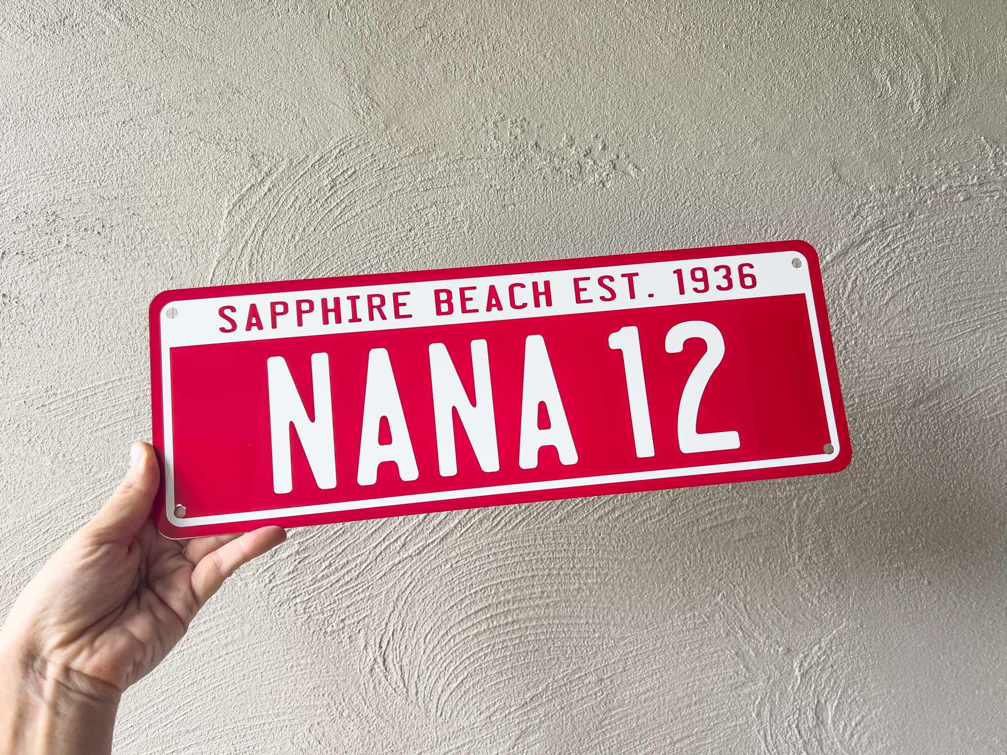 Red number plate style sign with white text that reads 'NANA 12' held against a light grey wall