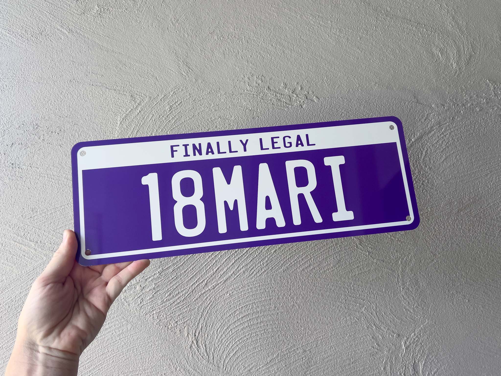 Hand holding a 18th Birthday purple license plate sign with text against a grey wall