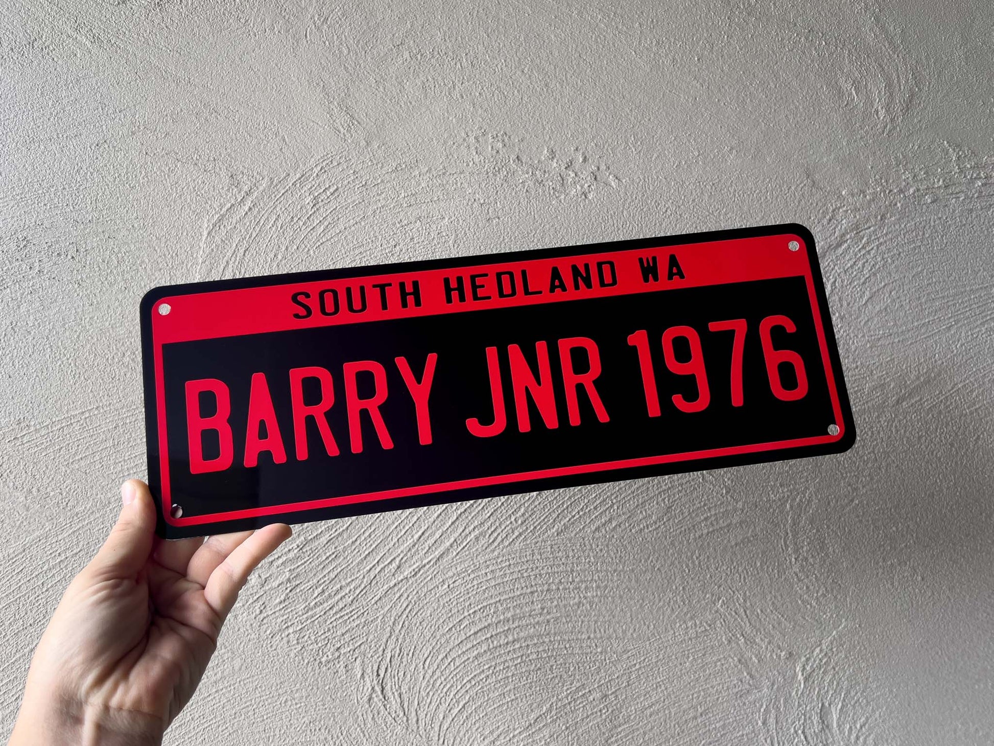 Person holding a red and black number plate sign with text against a light grey wall