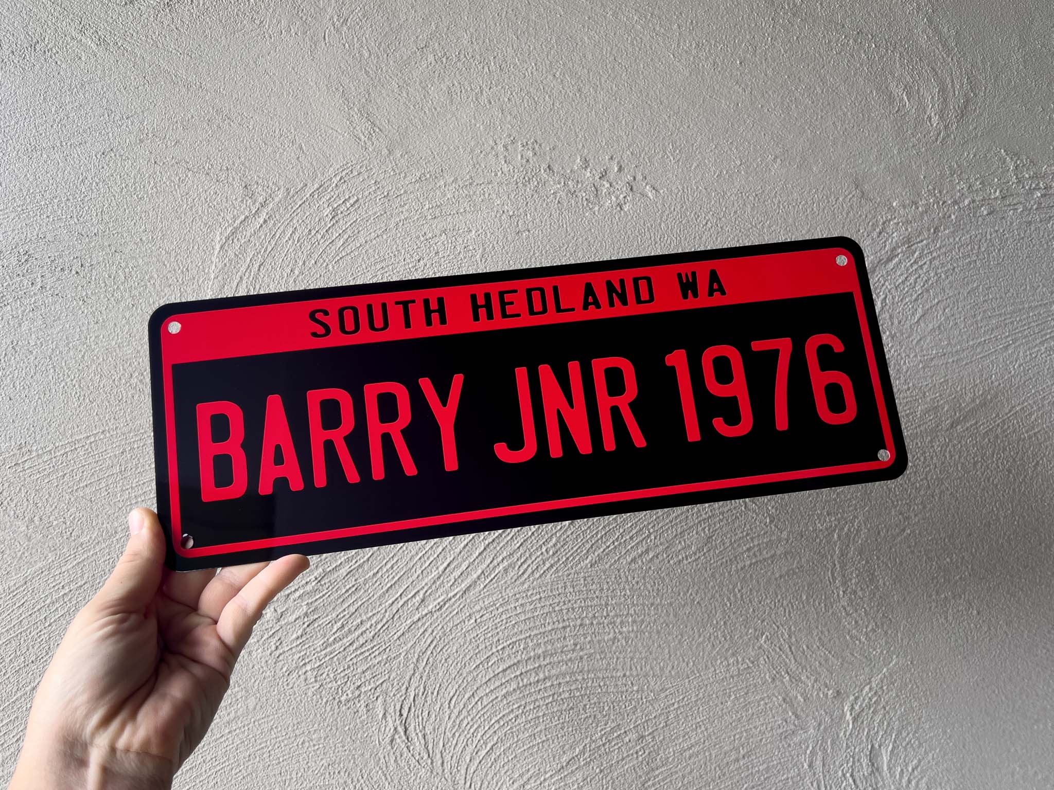 Person holding a red and black number plate sign with text against a light grey wall