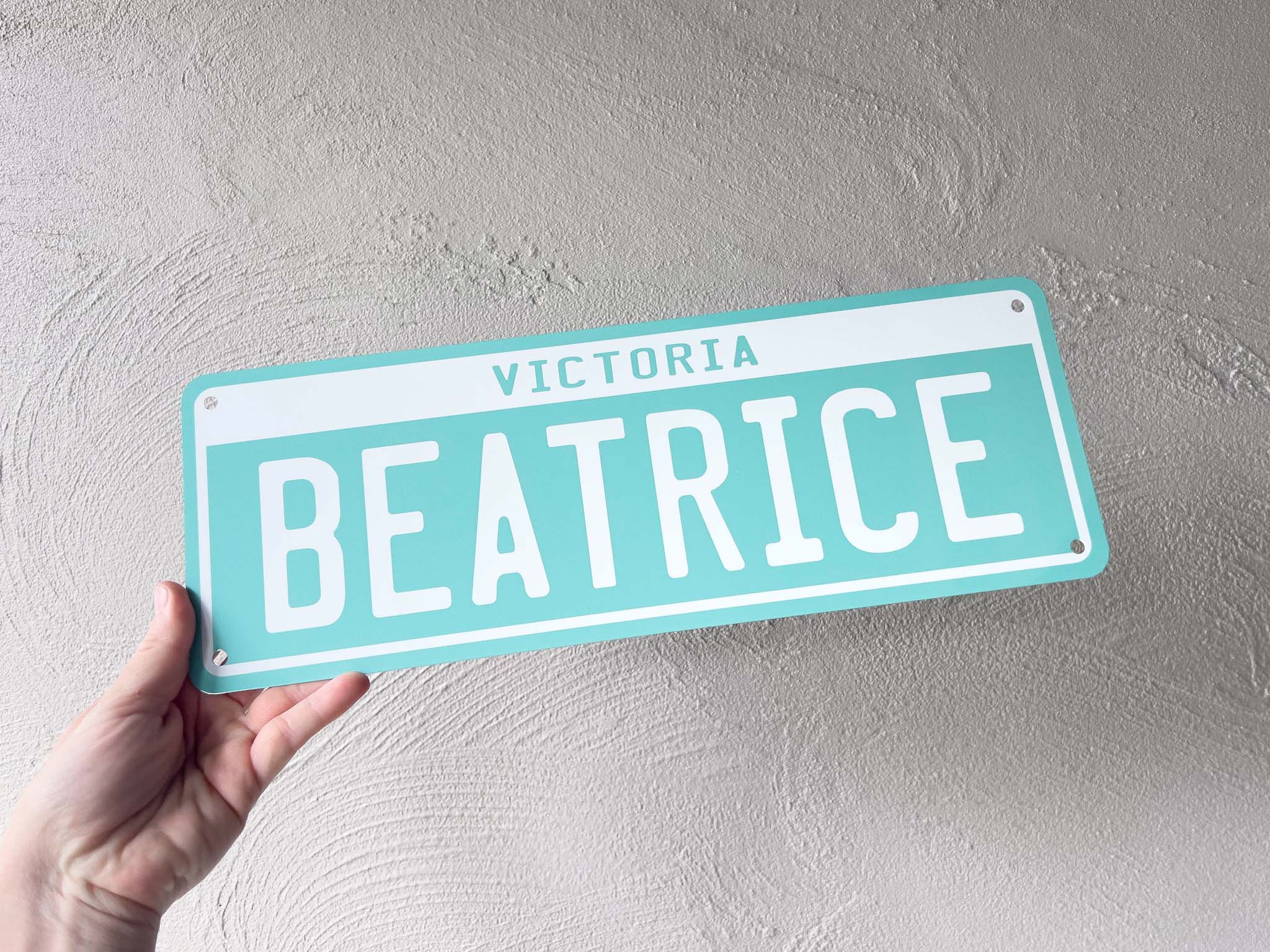 Hand holding a turquoise sign with 'BEATRICE' on a light gray background