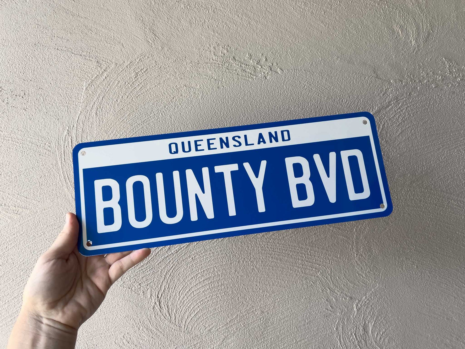 Hand holding a blue and white plate with 'Queensland Bounty BVD' text against a beige wall.