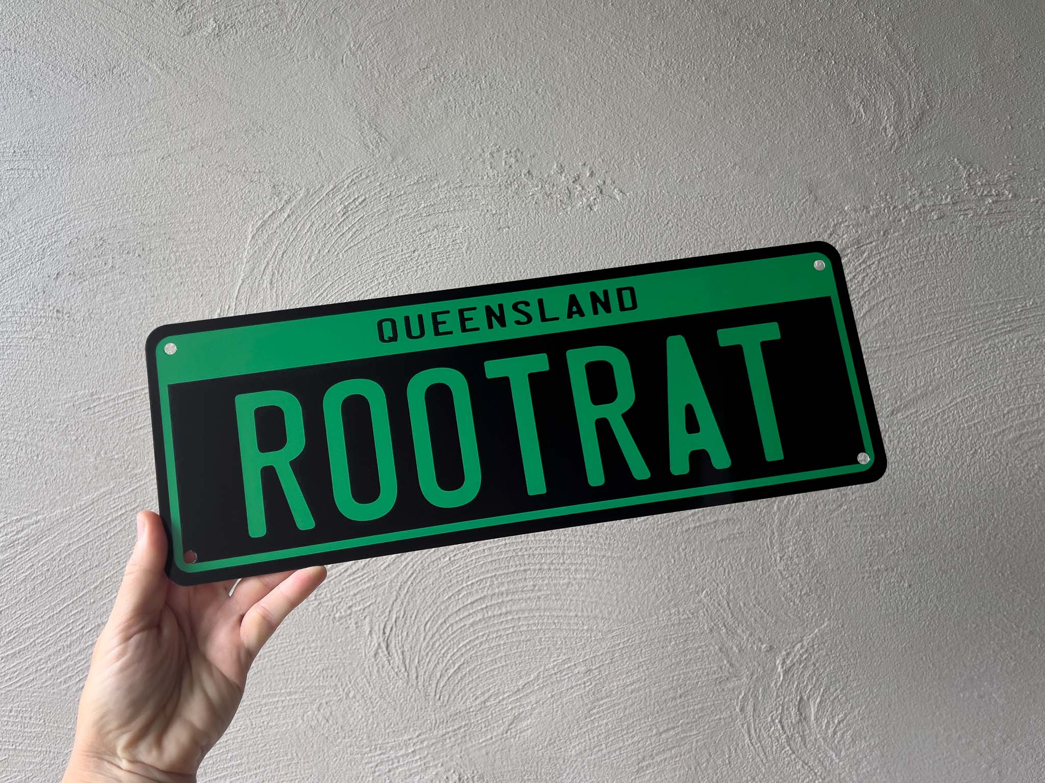 Hand holding a green and black sign with 'Queensland Rootrat' text against a light gray background