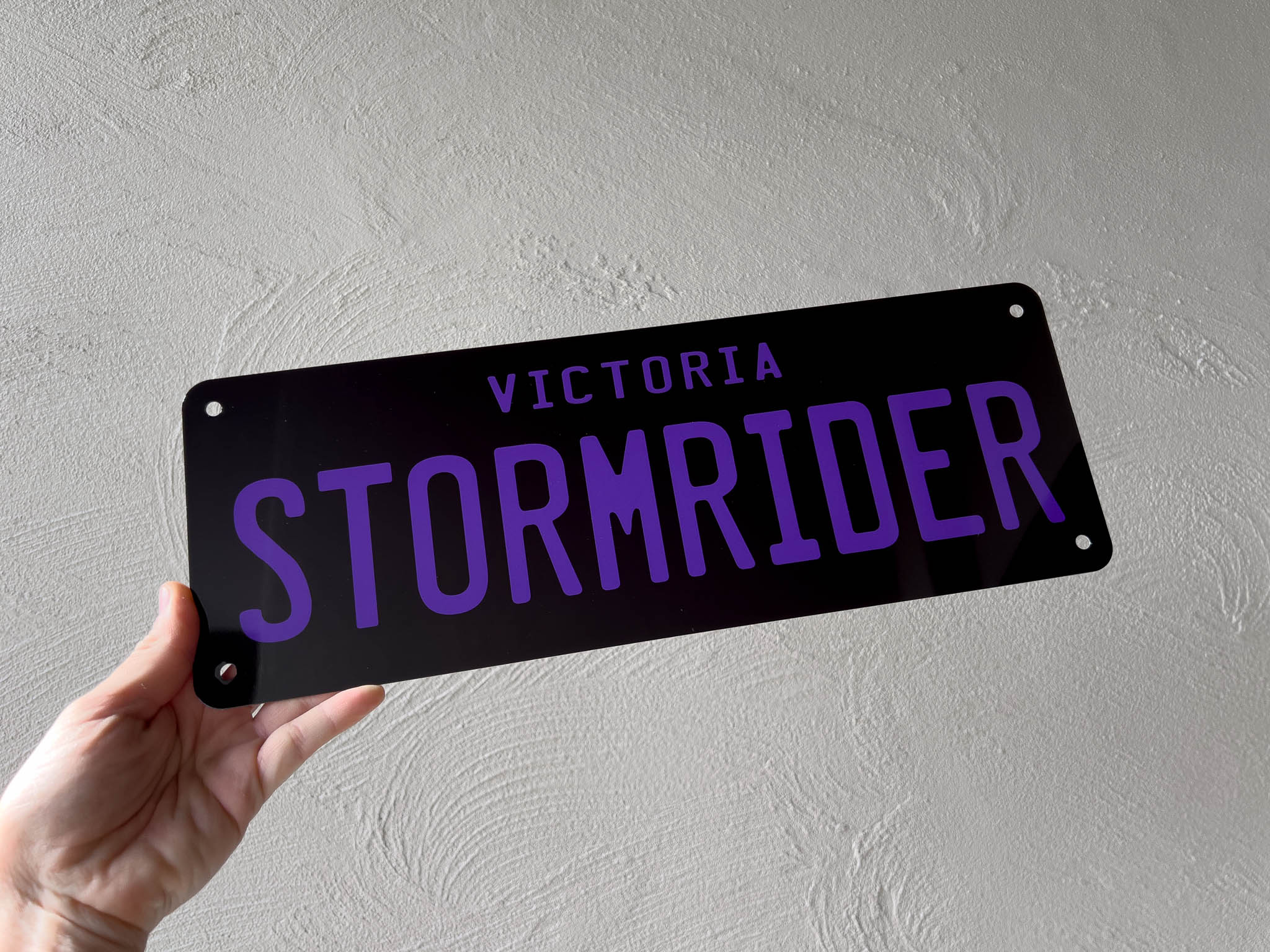 Hand holding a black sign with 'Victoria Stormrider' in purple text against a gray background