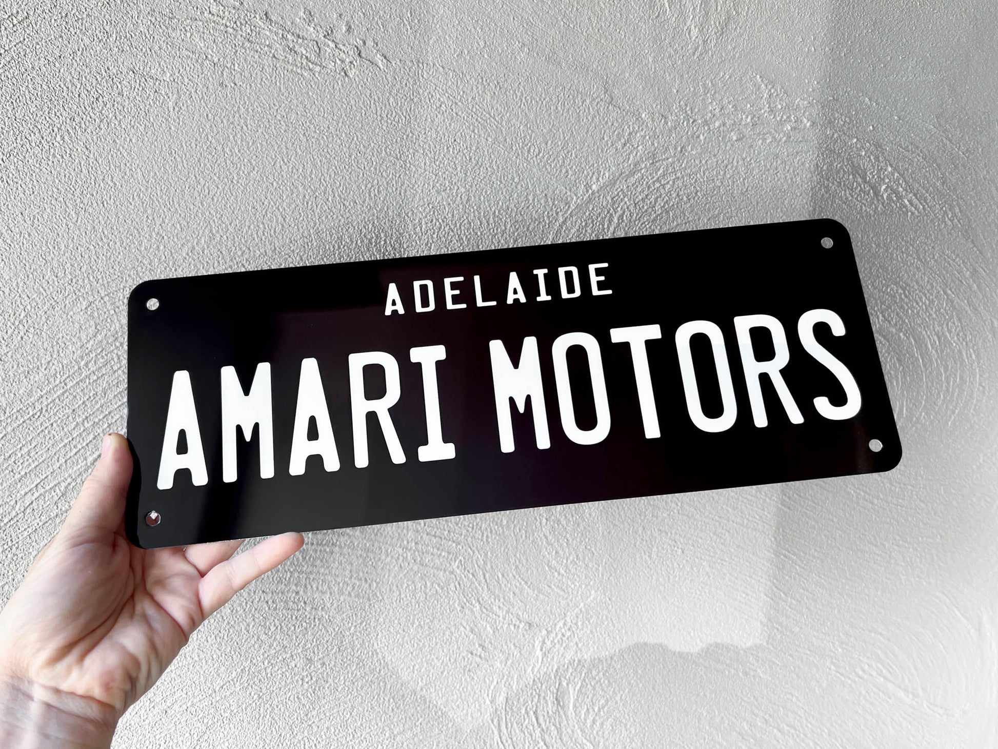 Black sign with 'ADELAIDE AMARI MOTORS' text held by a hand against a light gray background