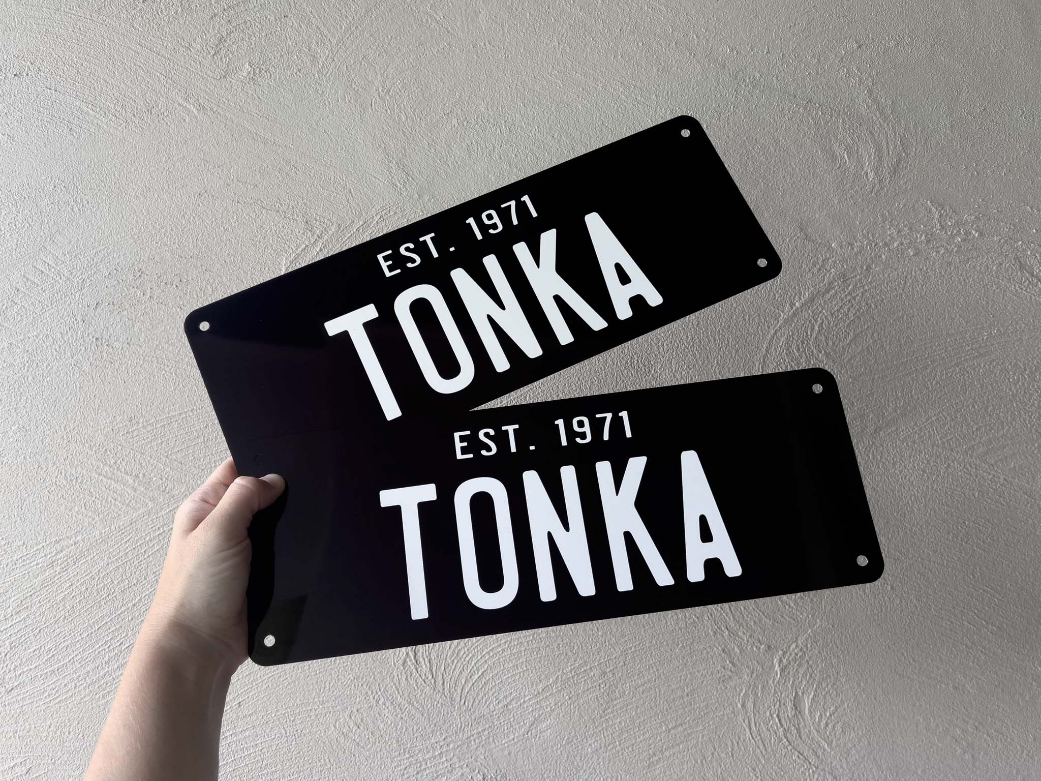 Two black metal signs with 'TONKA EST. 1971' text held against a light gray background.