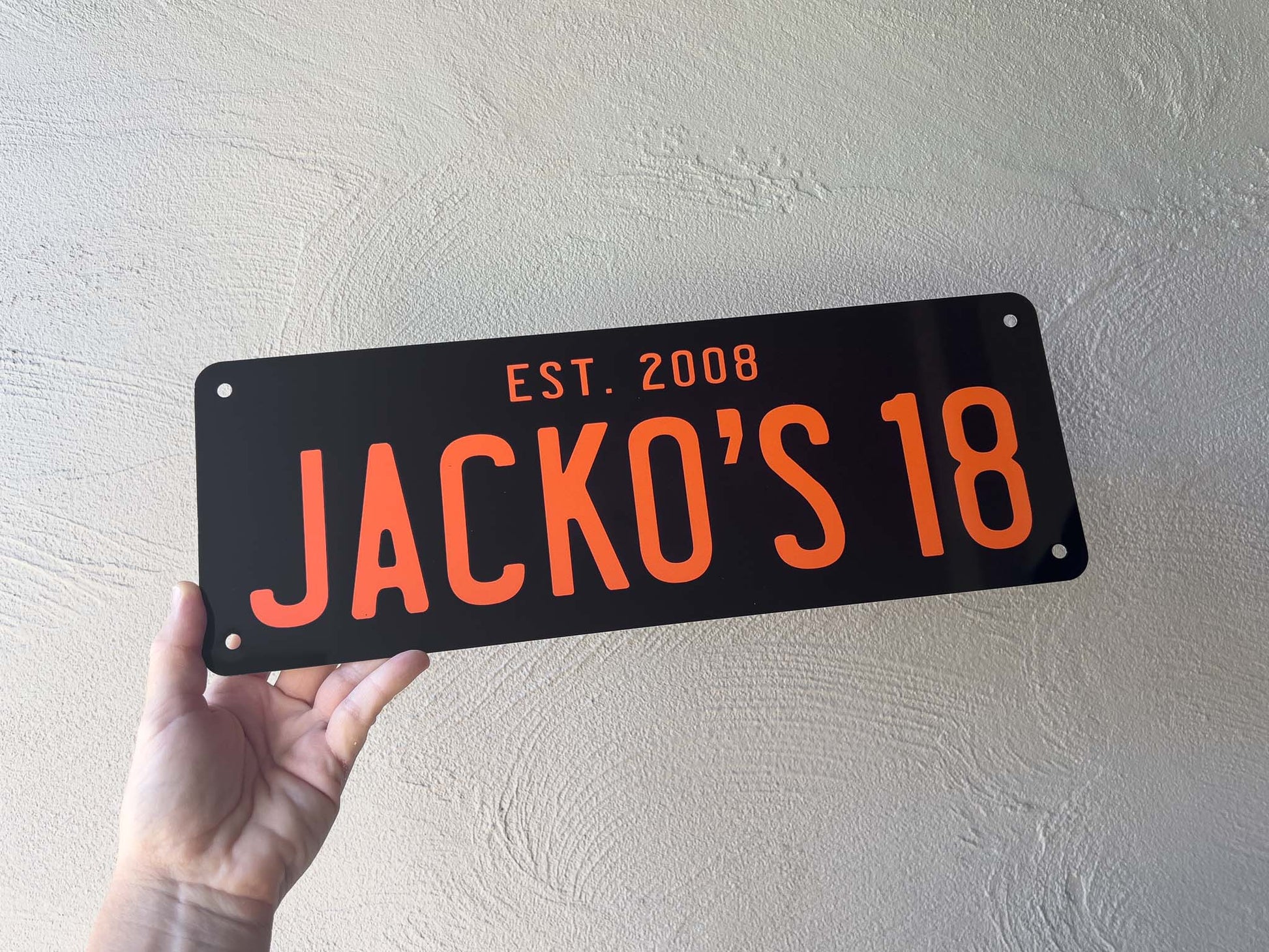 Hand holding a black plate with 'JACKO'S 18' in orange text against a light gray wall.