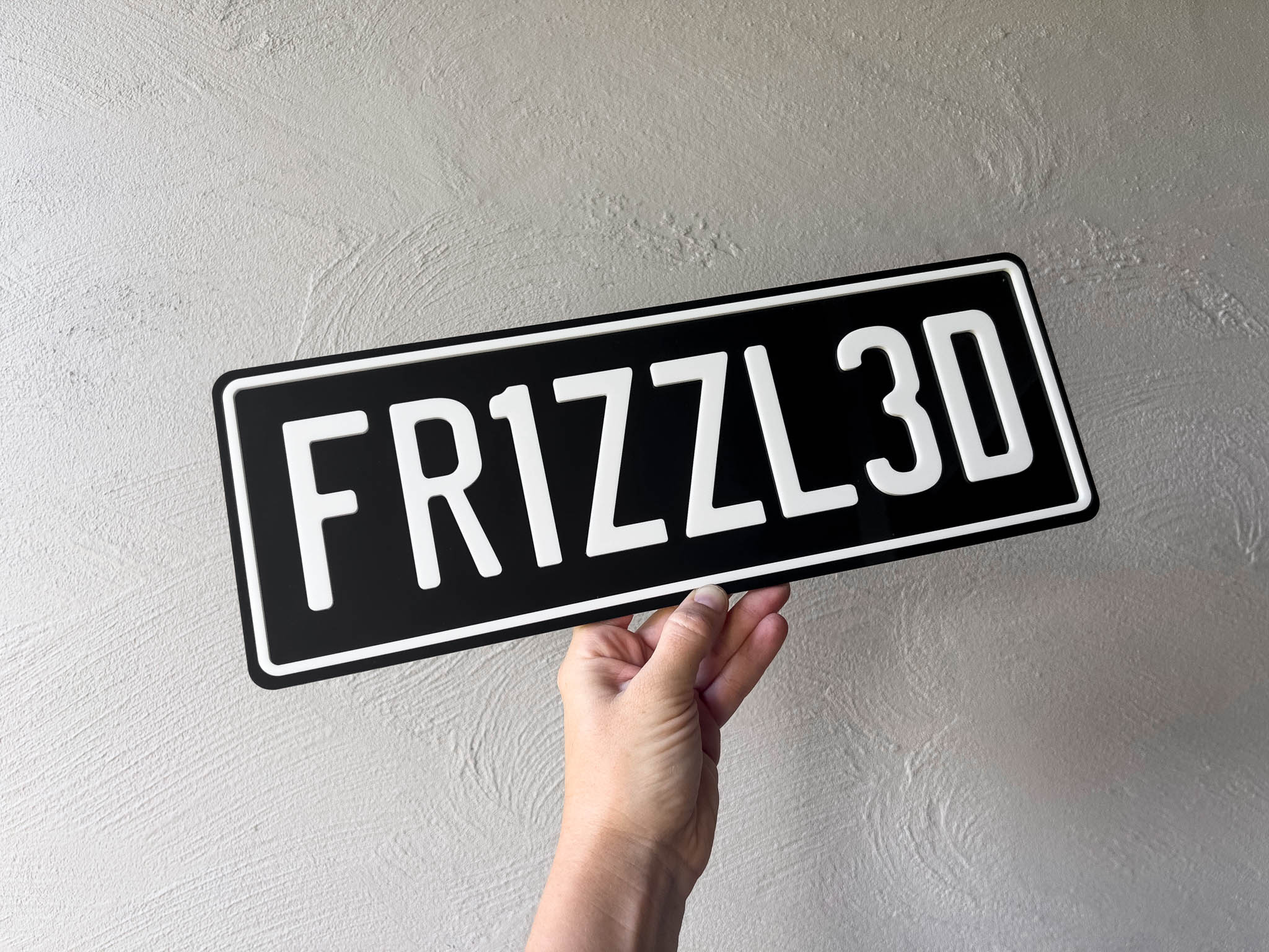 Hand holding a black and white number plate shaped sign in black and white that reads 'FR1ZZL3D'.