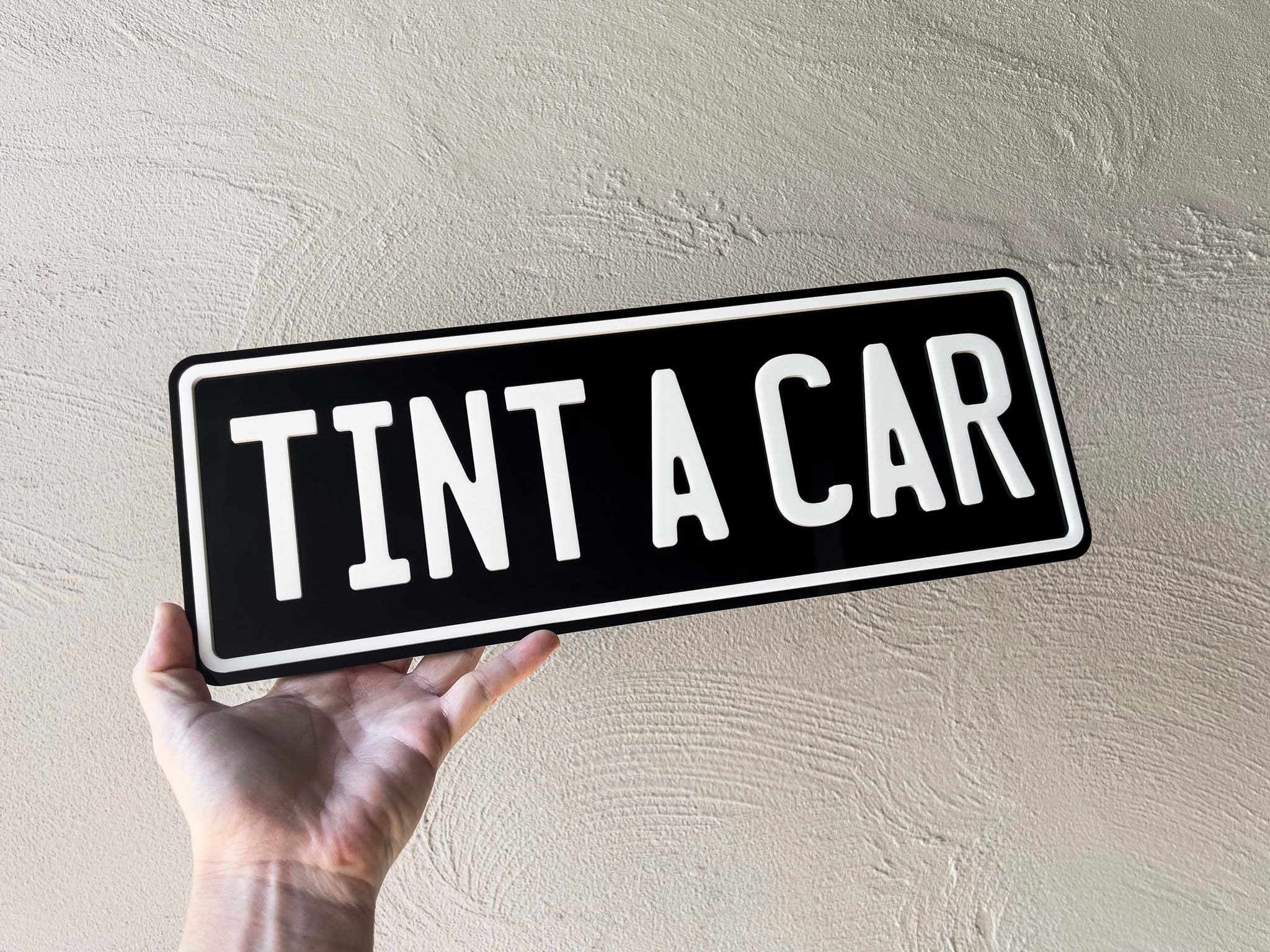 Hand holding a custom made 3D acrylic plate that says 'Tint A Car'.