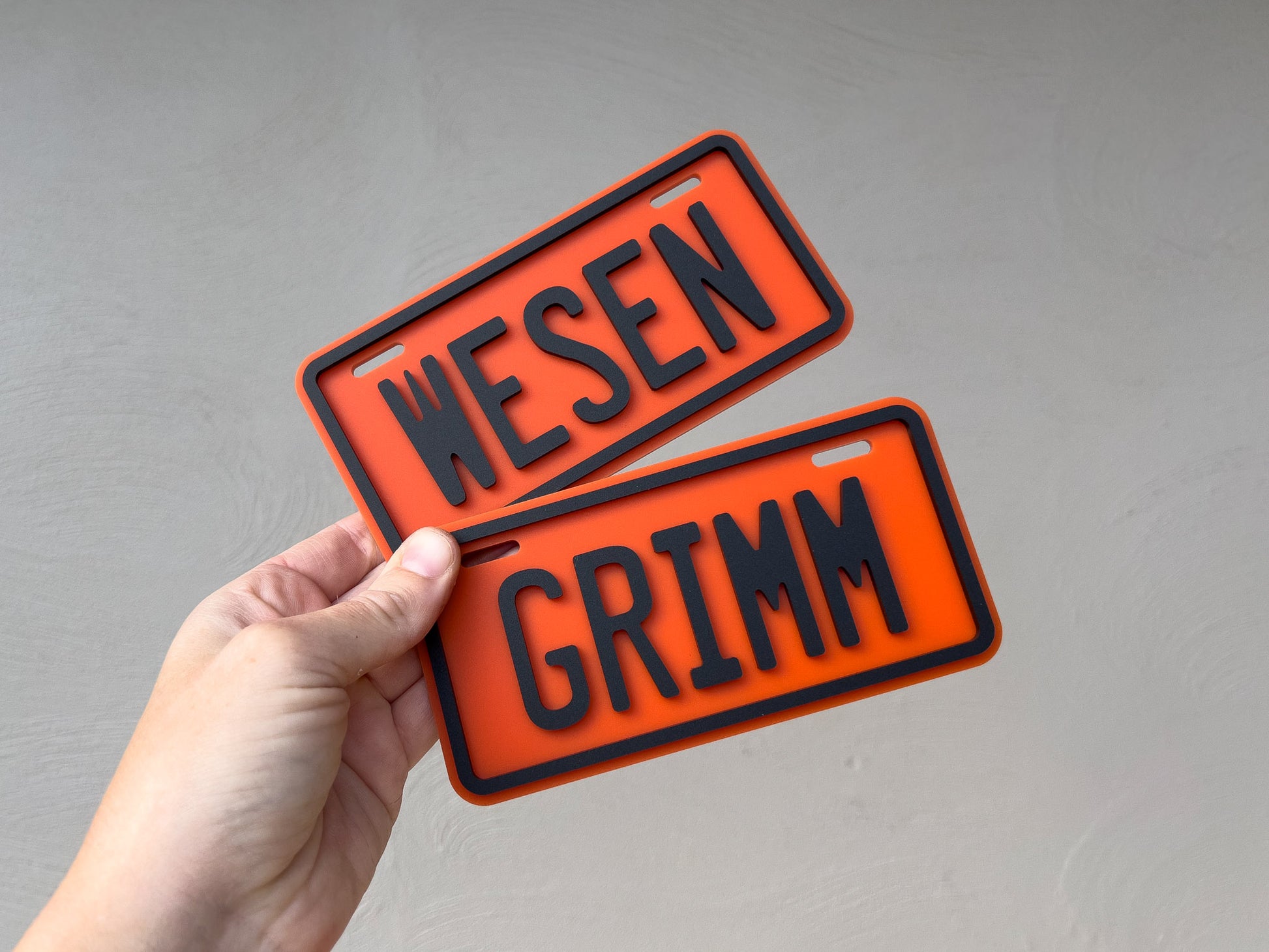 Two mini 3D acrylic number plates in orange with black 3D text that read 'WESEN' & 'GRIMM'.