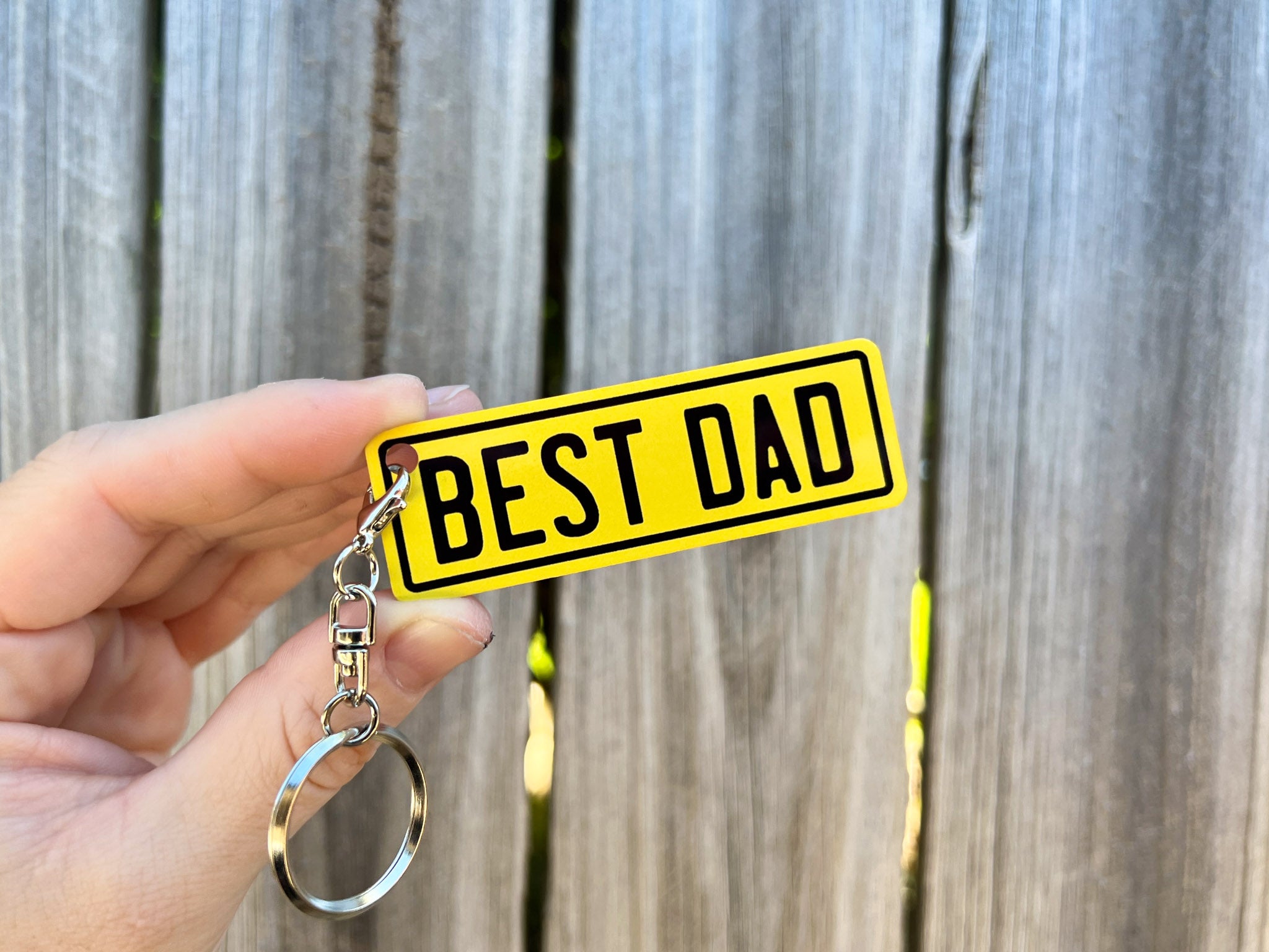 Keychain with 'BEST DAD' text held in front of a wooden fence