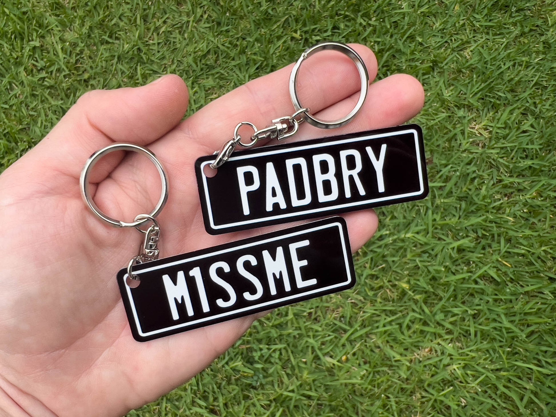 Two keychains with 'PADBRY' and 'M1SSME' text held in a hand against a grass background.