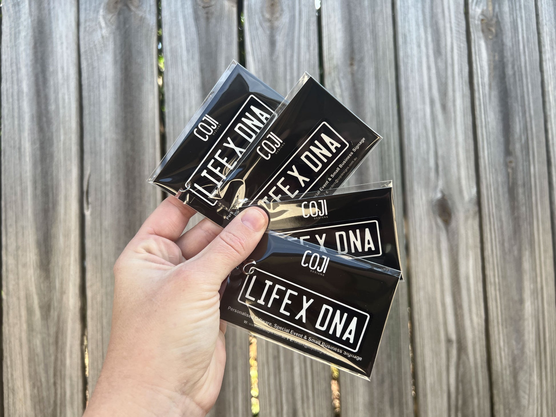 Hand holding three black and white custom printed 'LIFE X DNA' keychains against a wooden fence background.
