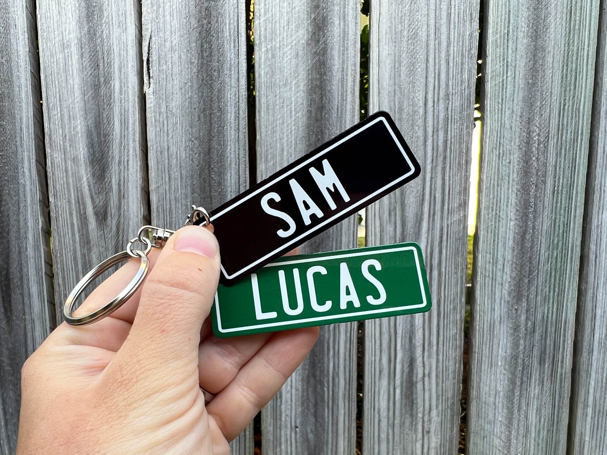 Two number plate style keychains with 'SAM' and 'LUCAS' text held against a wooden fence background.