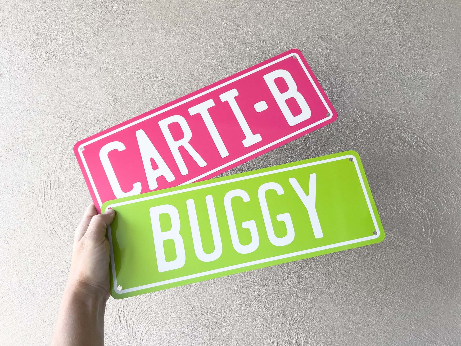 Two number plate-style signs with 'CARTI-B' and 'BUGGY' text held against a light grey background.