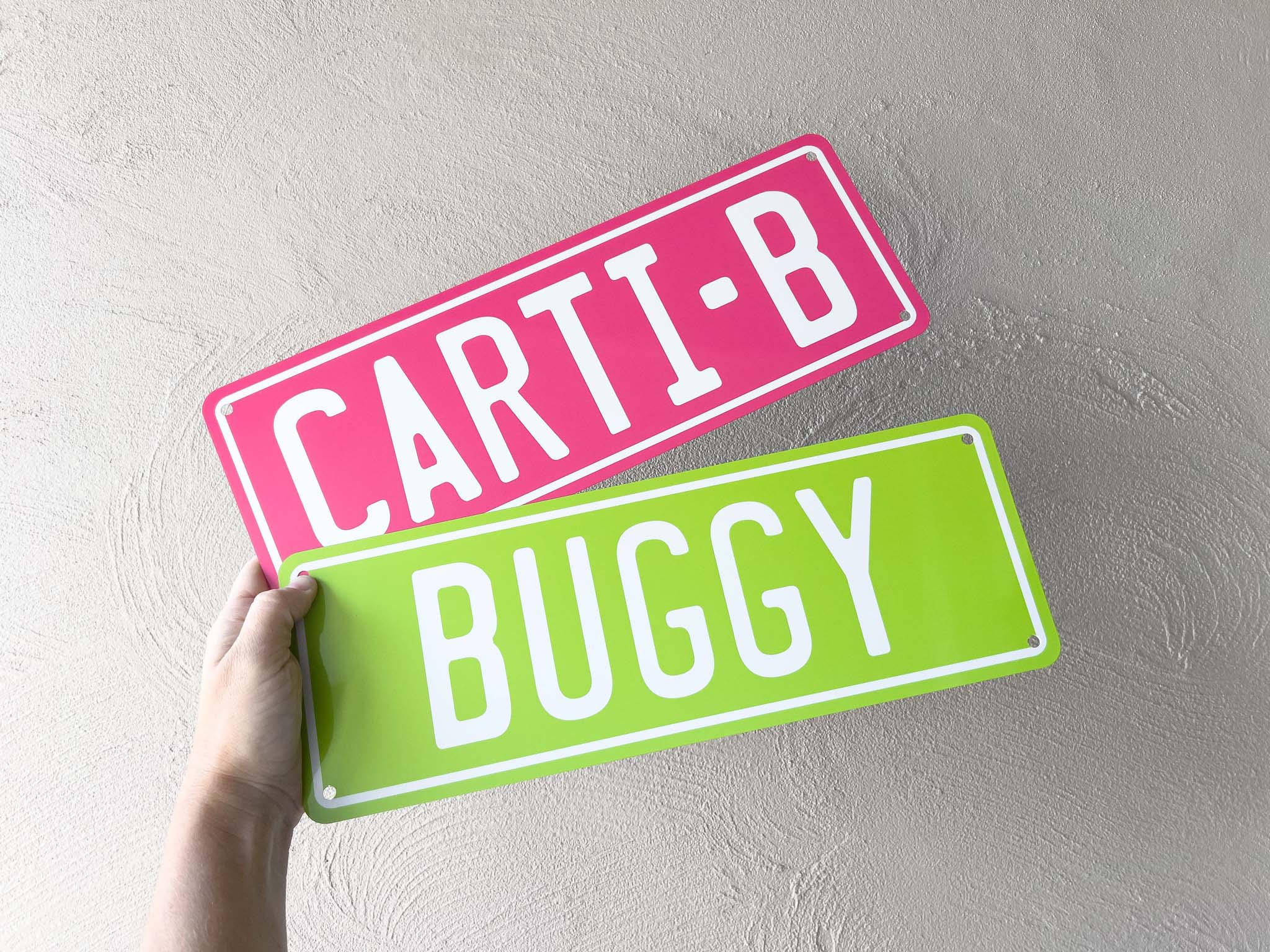 Two number plate-style signs with 'CARTI-B' and 'BUGGY' text held against a light grey background.