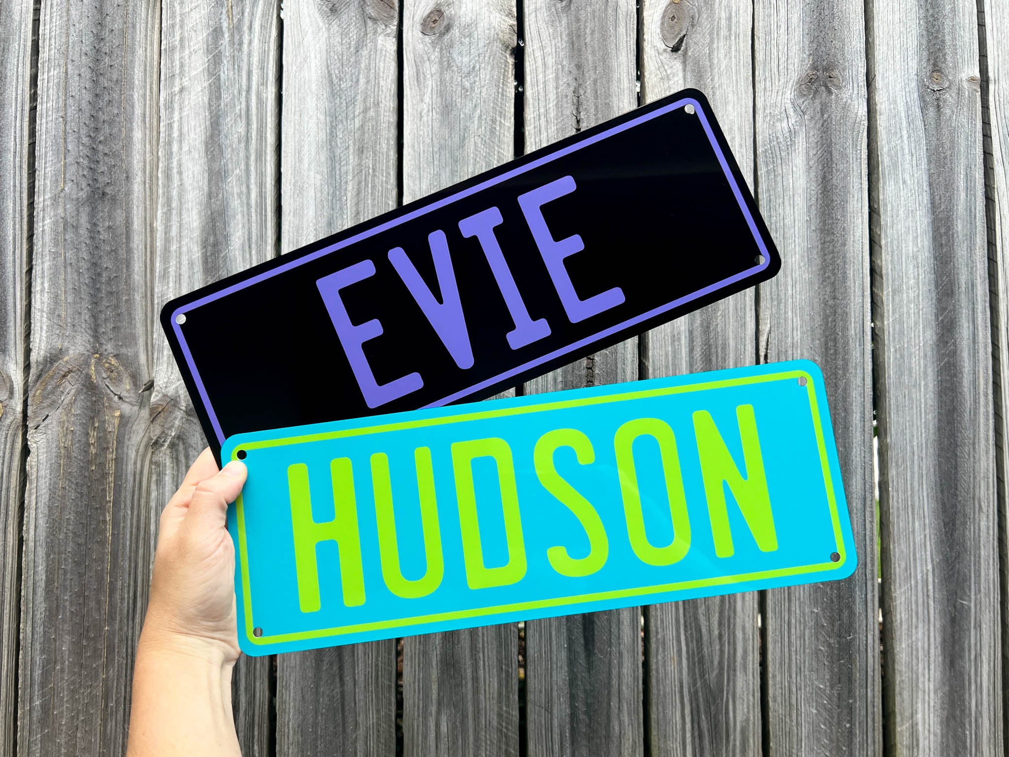 Two novelty number plate nameplates, one with 'EVIE' and another with 'HUDSON', held against a wooden background.
