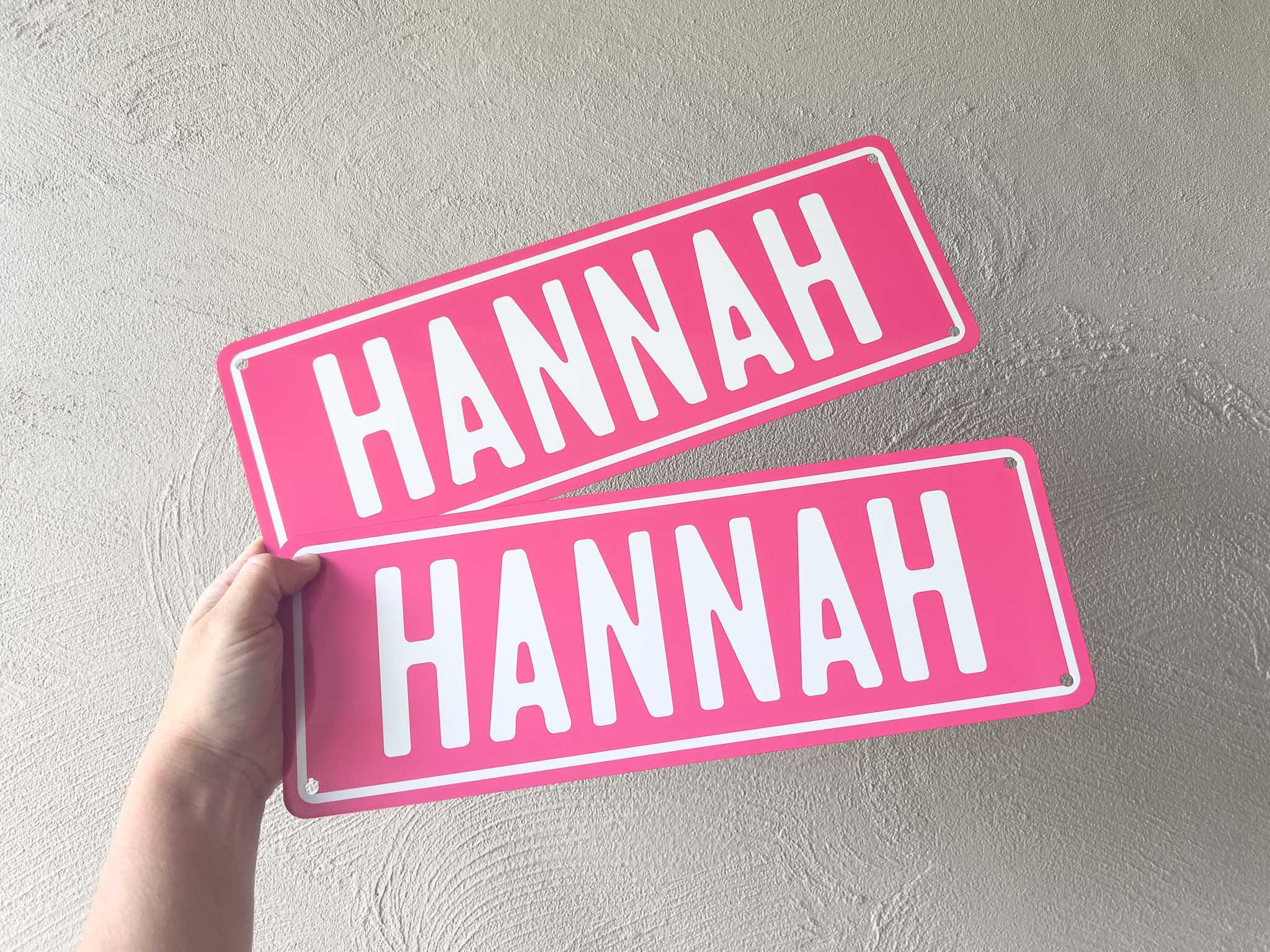 Two pink and white rectangular aluminium plates with 'HANNAH' printed on them, held against a light grey background.
