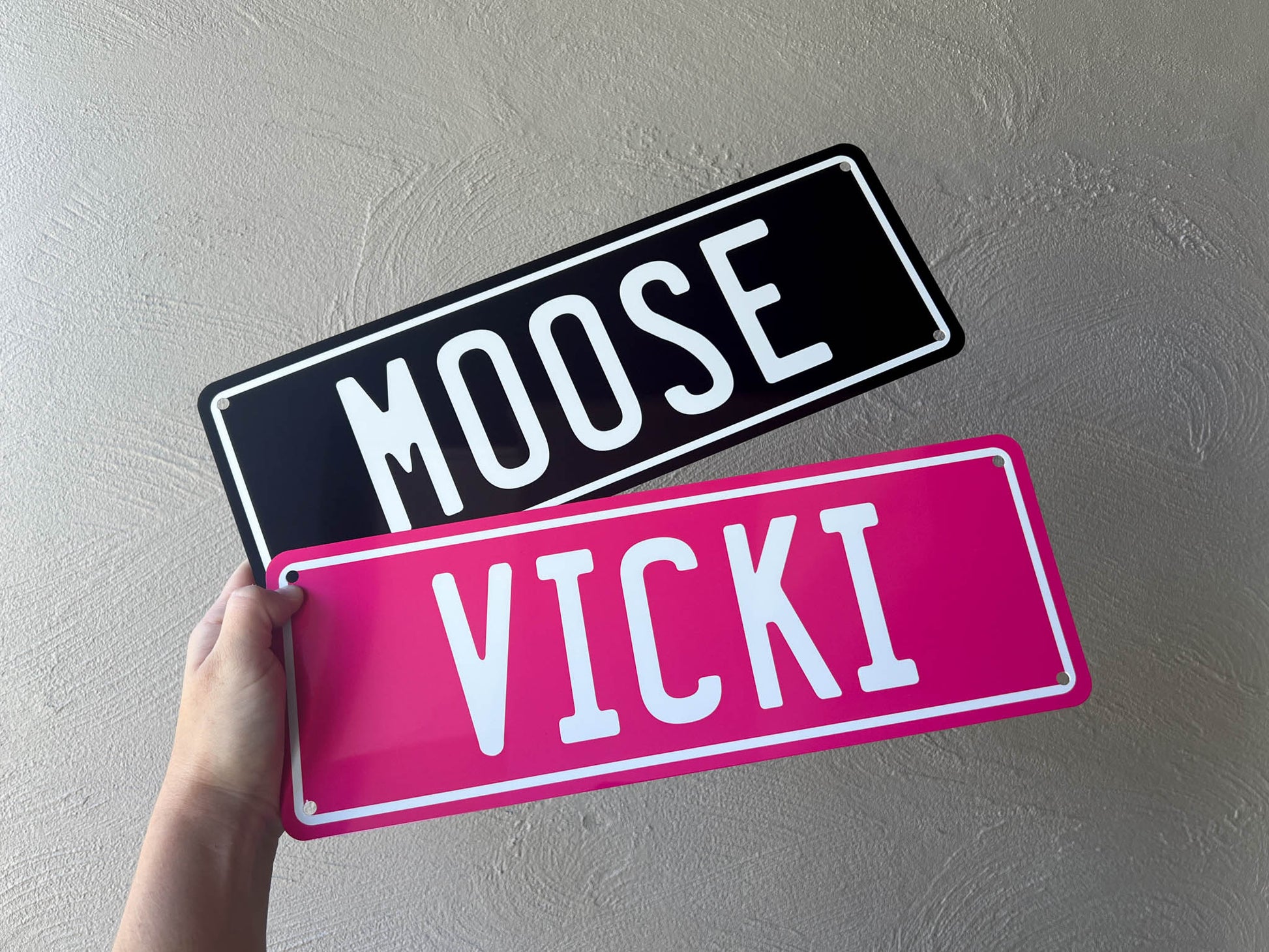 Two number plate nameplates, one black with 'MOOSE' and one pink with 'VICKI', held against a plain background.