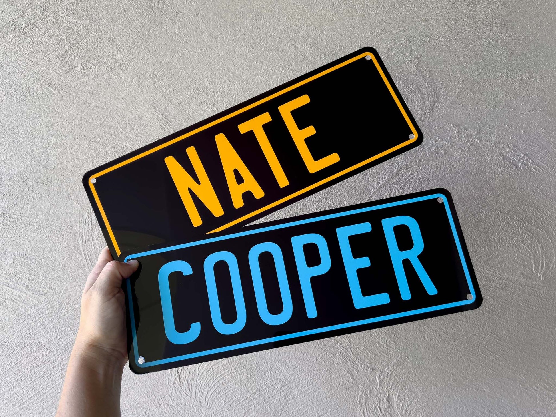 Two metal nameplates held against a light gray wall, one with 'NATE' in yellow and the other with 'COOPER' in blue.
