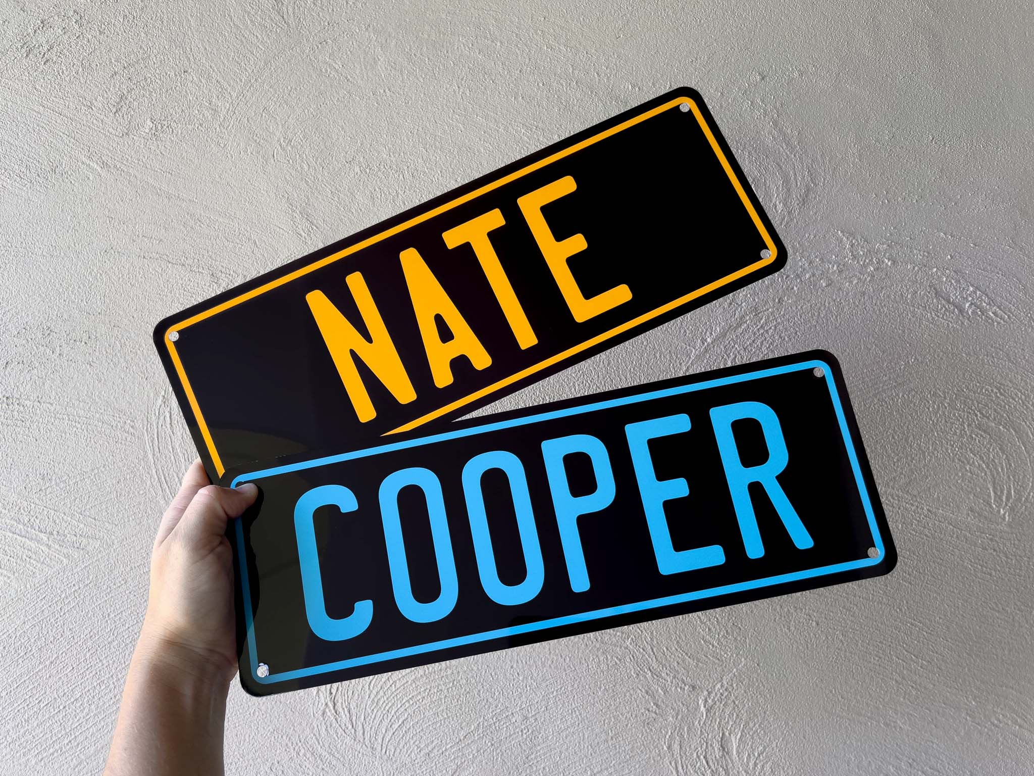 Two metal nameplates held against a light gray wall, one with 'NATE' in yellow and the other with 'COOPER' in blue.