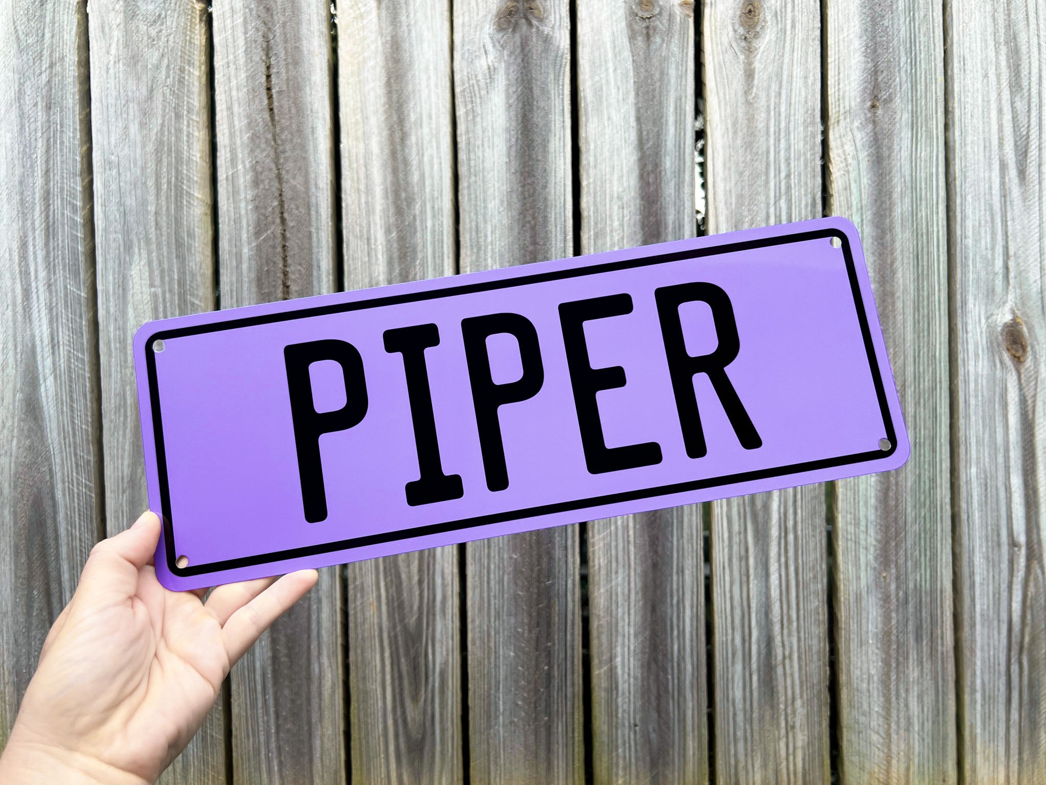 Hand holding a purple sign with 'PIPER' text against a wooden fence background
