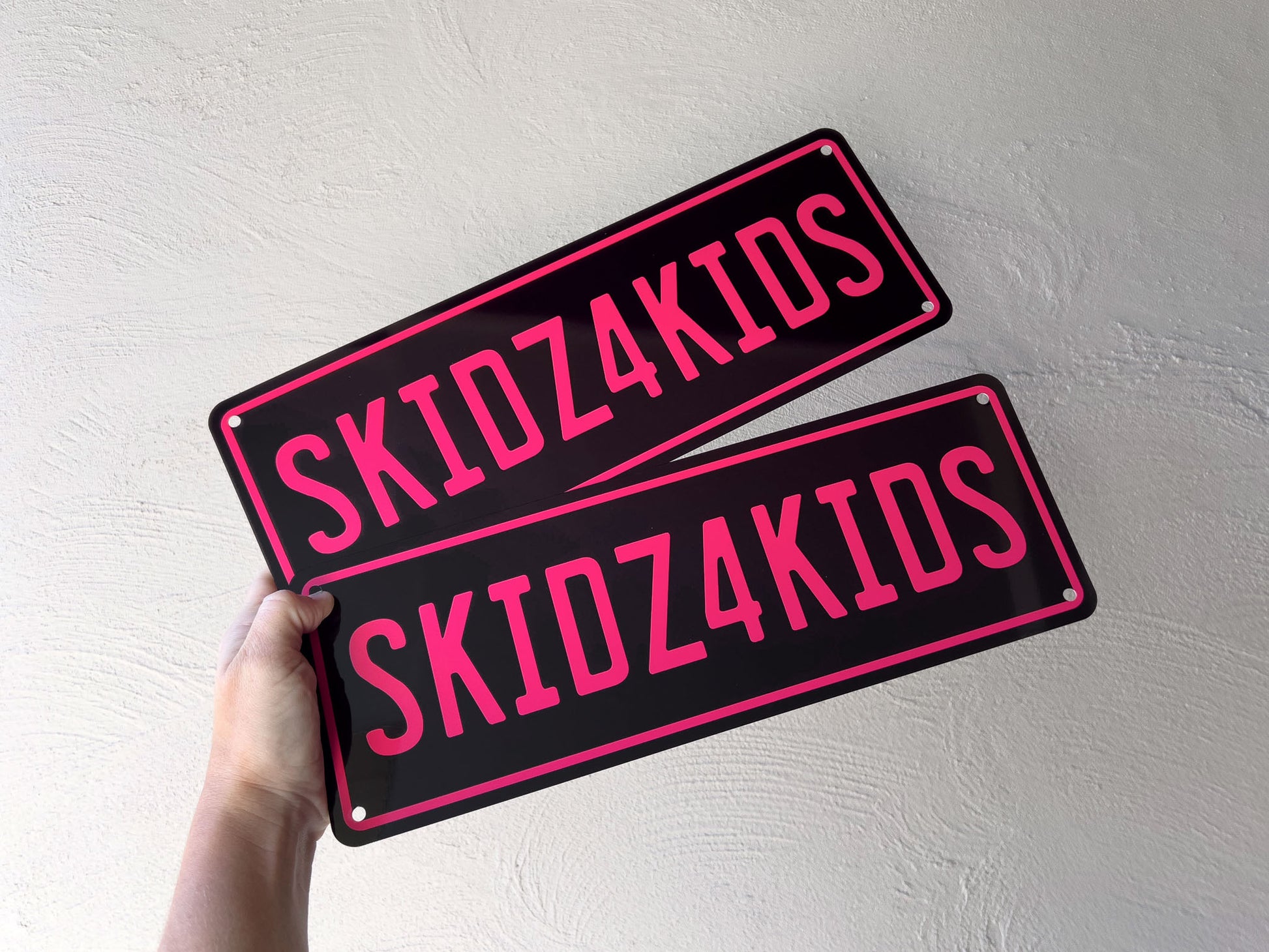 Two custom printed black and magenta 'SKIDZ4KIDS' plates held by a hand against a light background