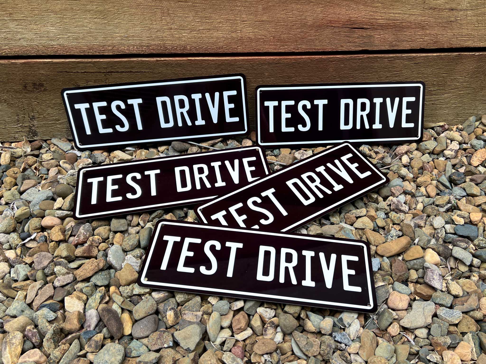 Multiple 'TEST DRIVE' metal black and white plates on a pebbly surface