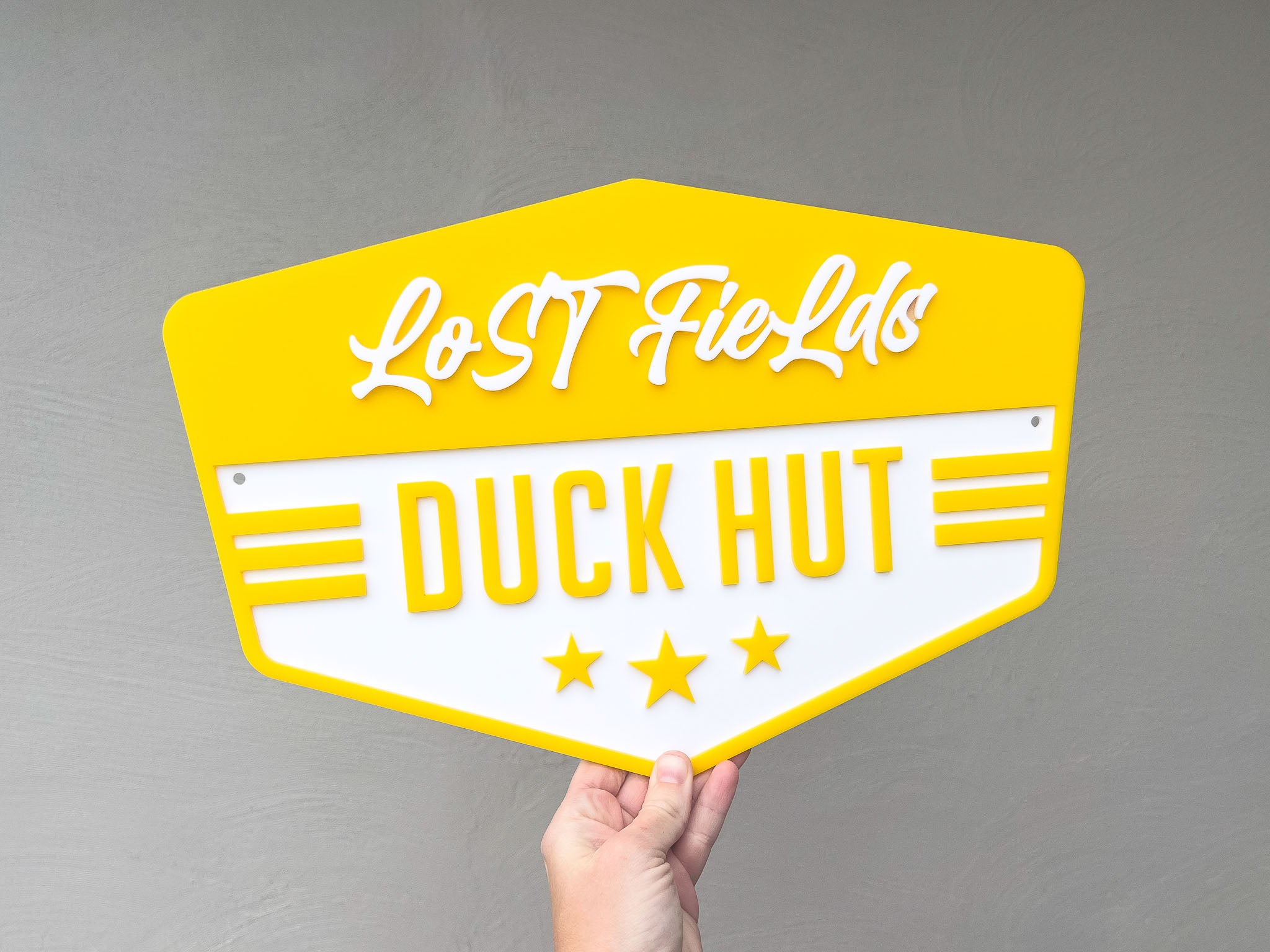 Badge shape sign in bright sunshine yellow with 'Lost Fields Duck Hut' text held against a grey background