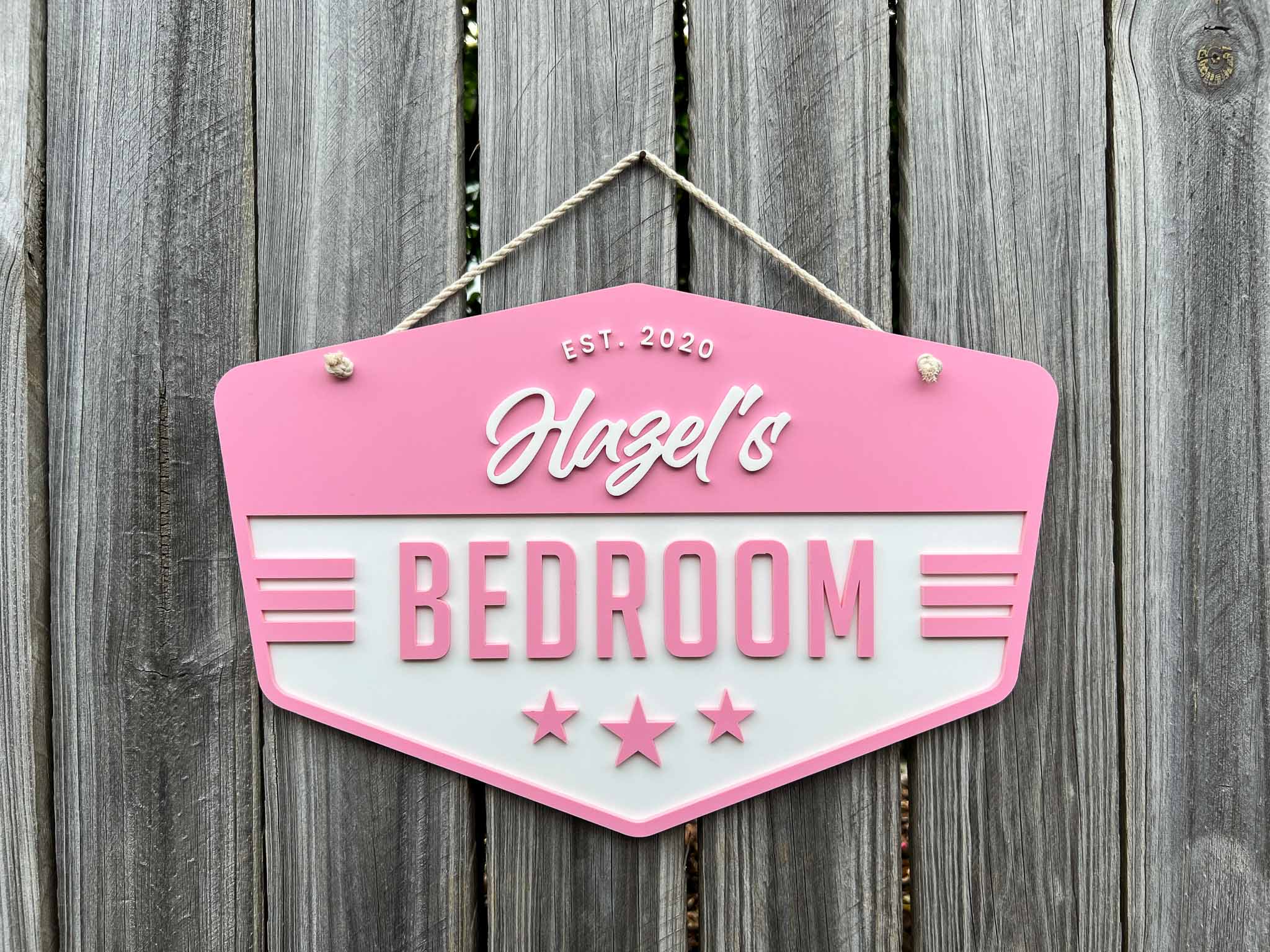 Pink and white sign with 'Hazel's Bedroom' text hanging with a natural rope on a wooden fence.