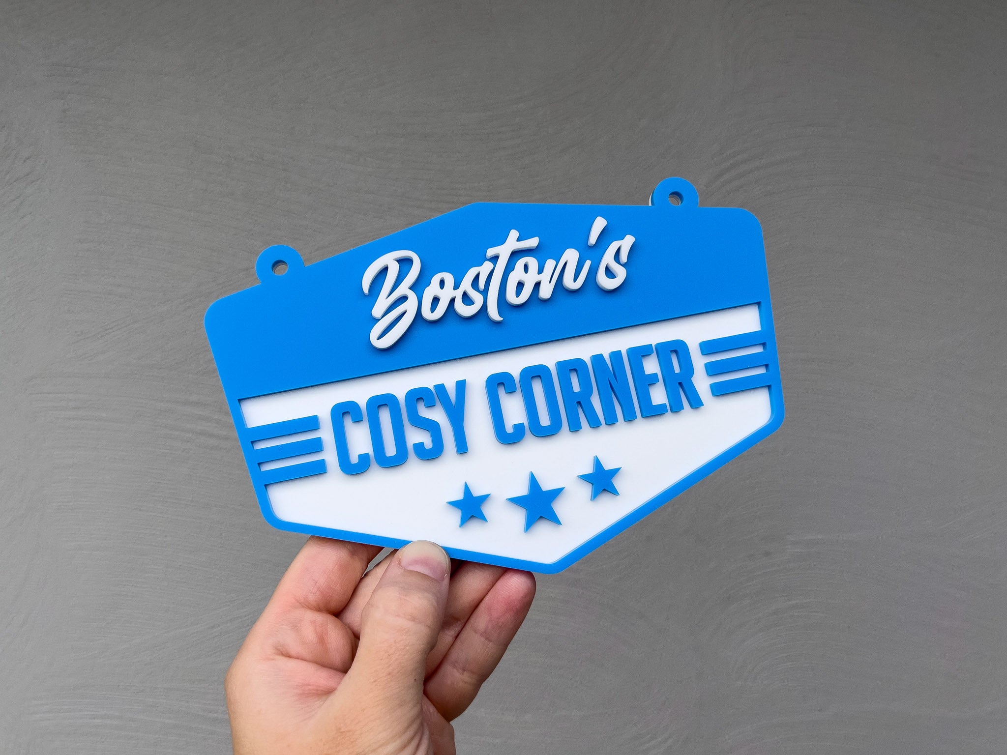 Hand holding a blue and white sign with 'Boston's Cosy Corner' text against a grey background | Coji Designs