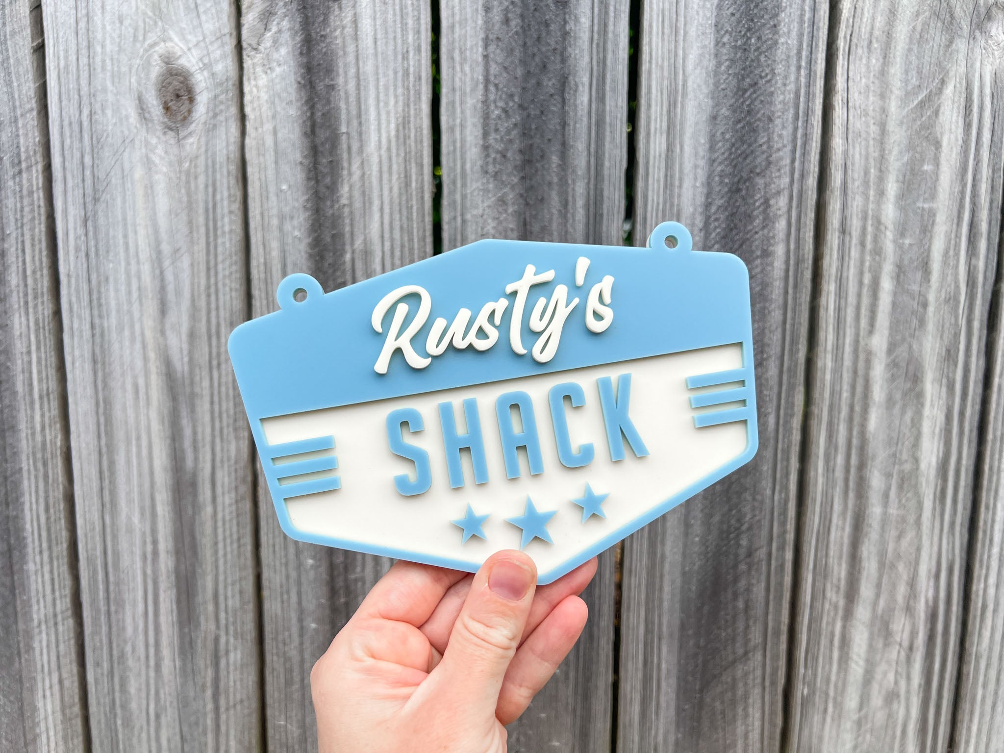 Hand holding a sign with 'Rusty's Shack' against a wooden background | Coji Designs