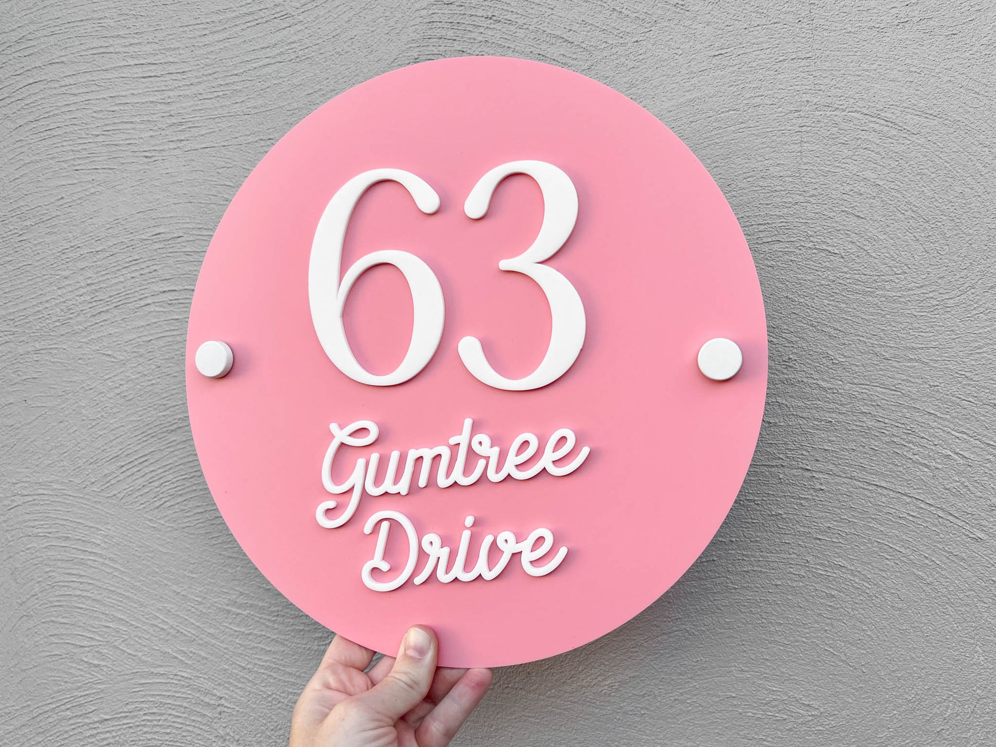 Candy pink melon and white colour round plaque with a street address that says '63 Gumtree Drive' with white wall mounts.