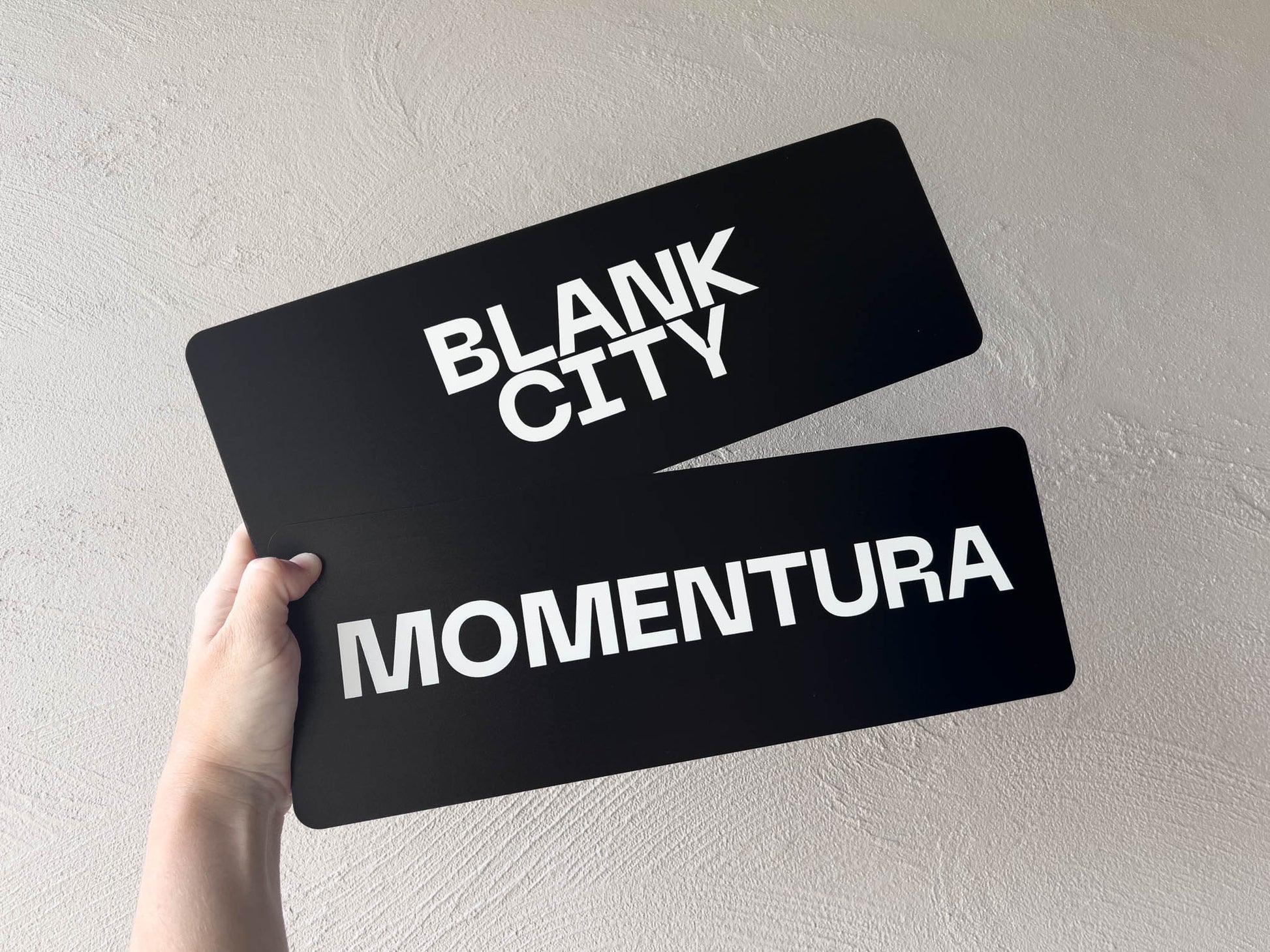 Two black and white flexi plates with 'Blank City' and 'Momentura' text held against a light gray background.