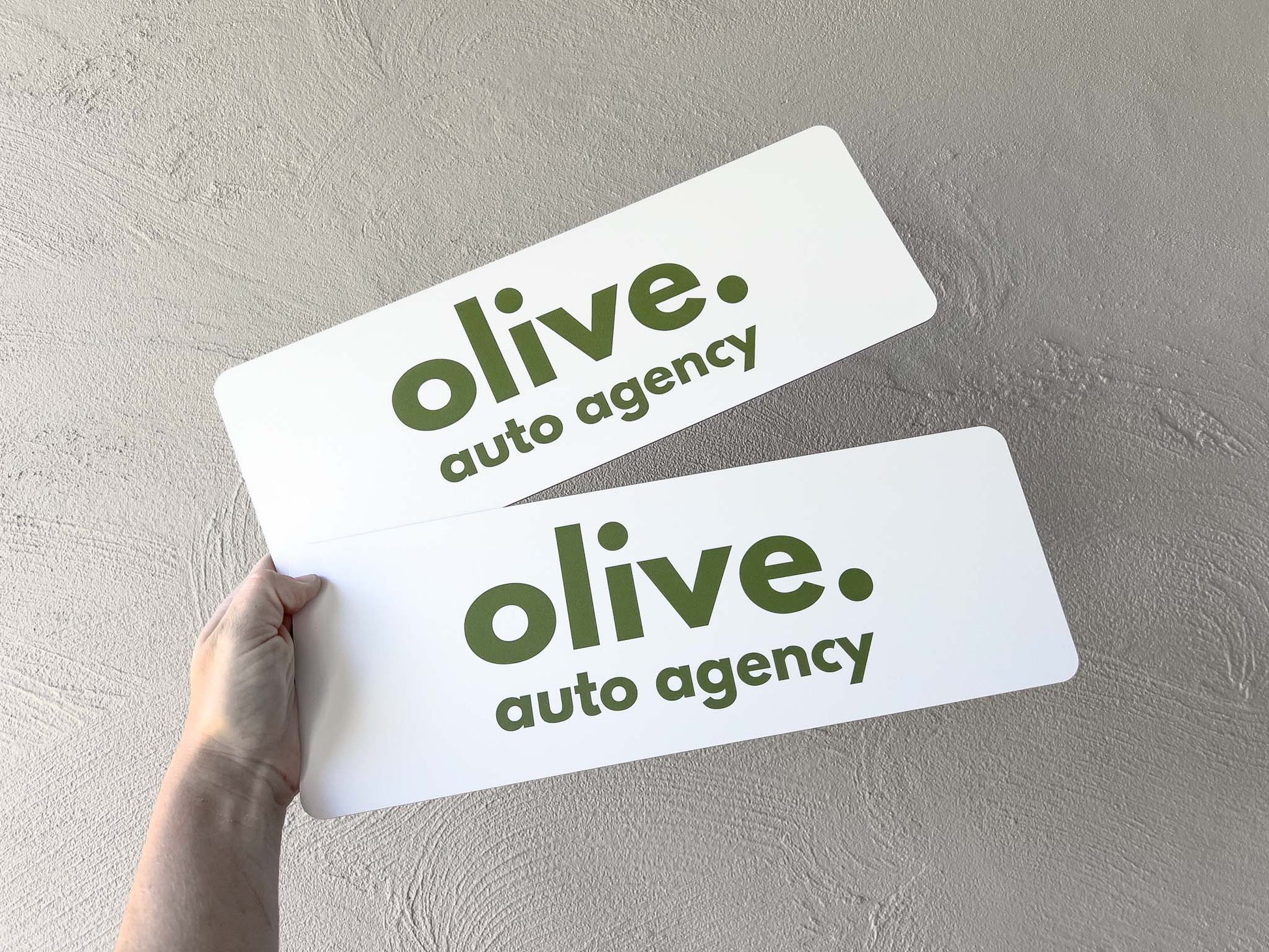 Two printed logo plates with 'olive auto agency' logo on a gray background