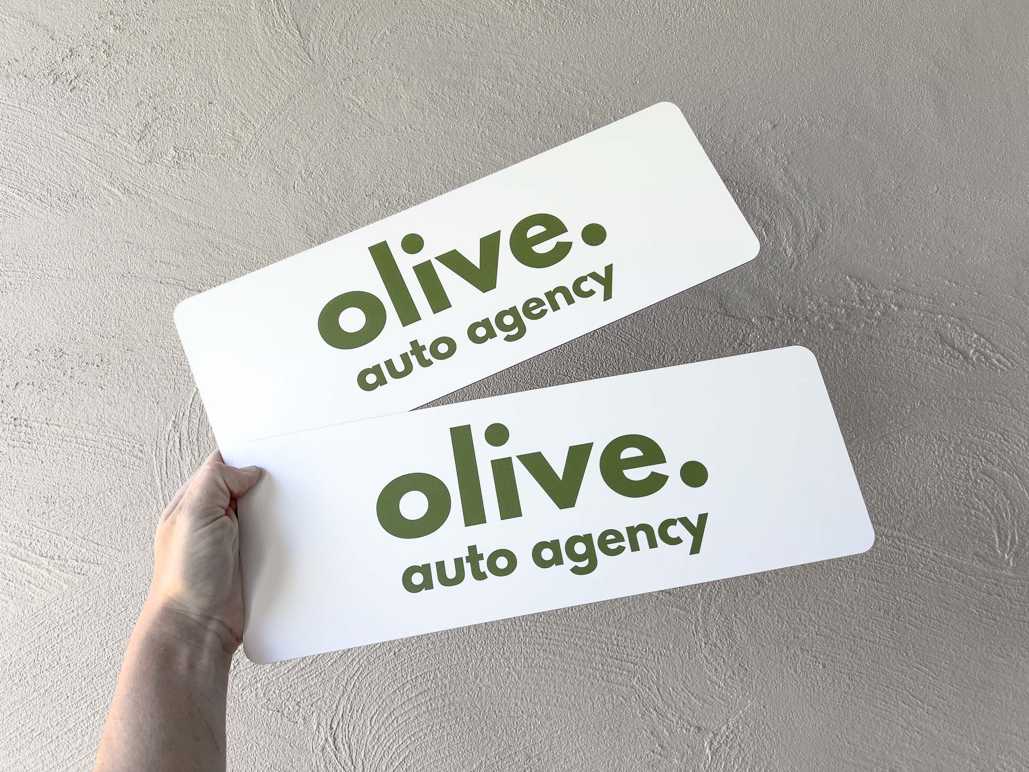 Two printed logo plates with 'olive auto agency' logo on a gray background