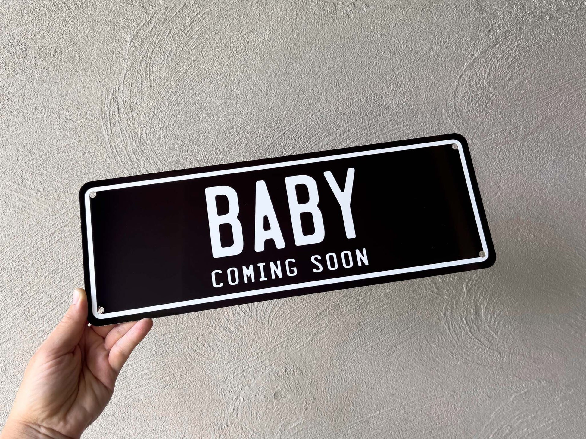 Hand holding a black plate with 'BABY COMING SOON' text against a light gray wall.
