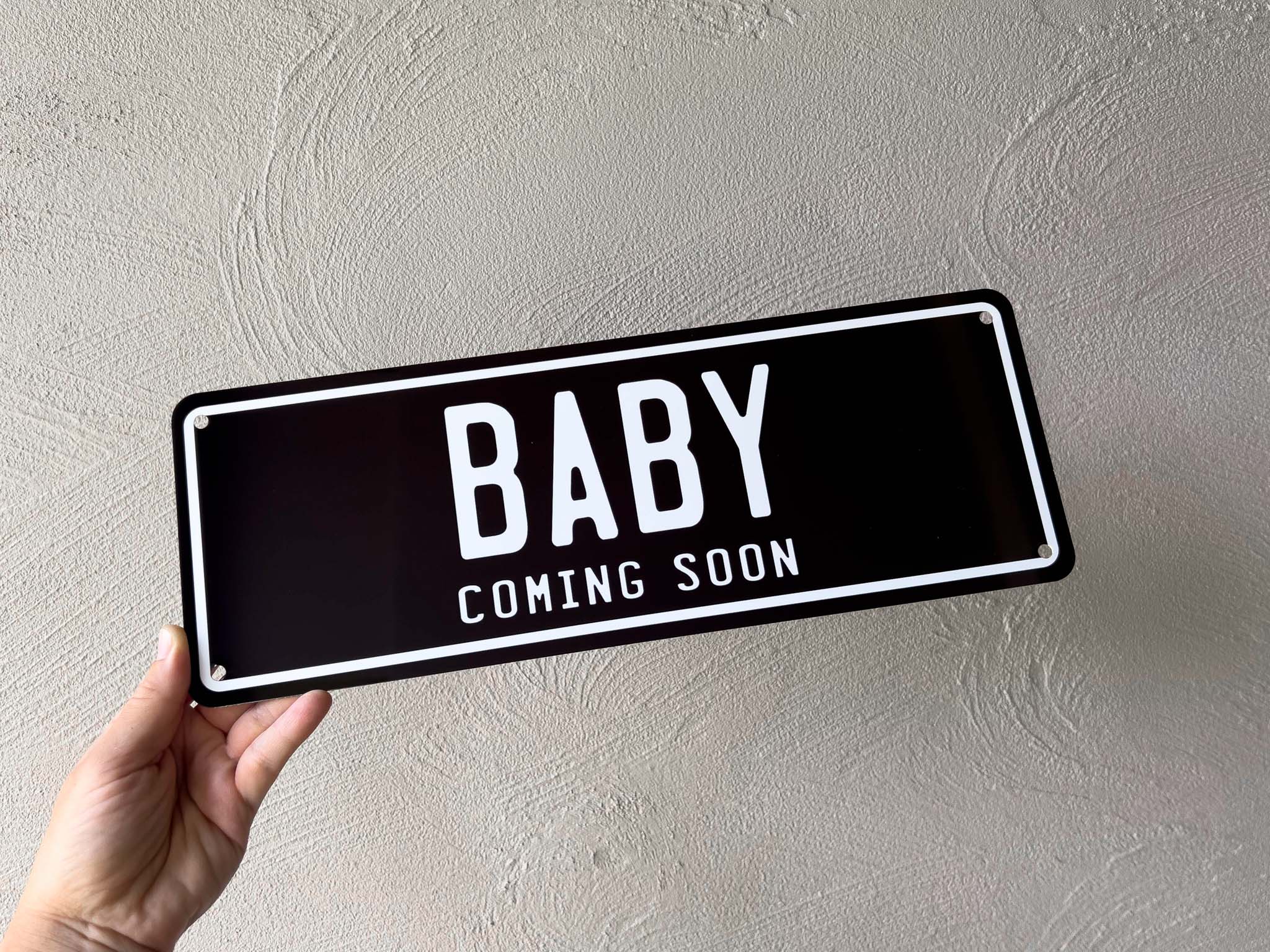 Hand holding a black plate with 'BABY COMING SOON' text against a light gray wall.