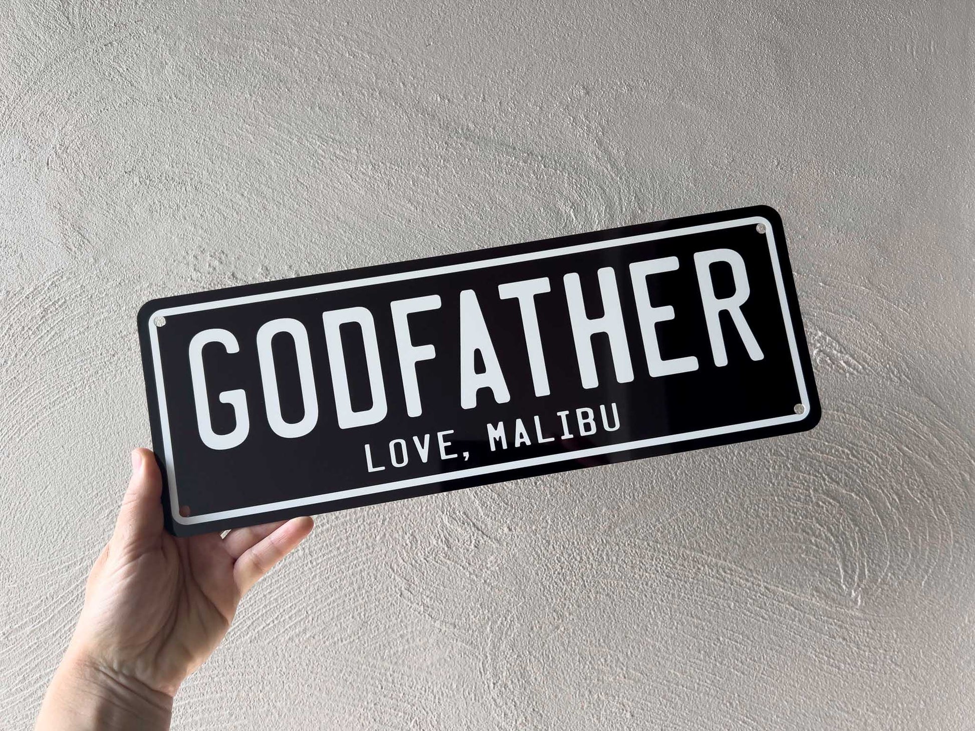 Hand holding a black and white plate with 'GODFATHER' and 'LOVE, MALIBU' text against a light grey background.