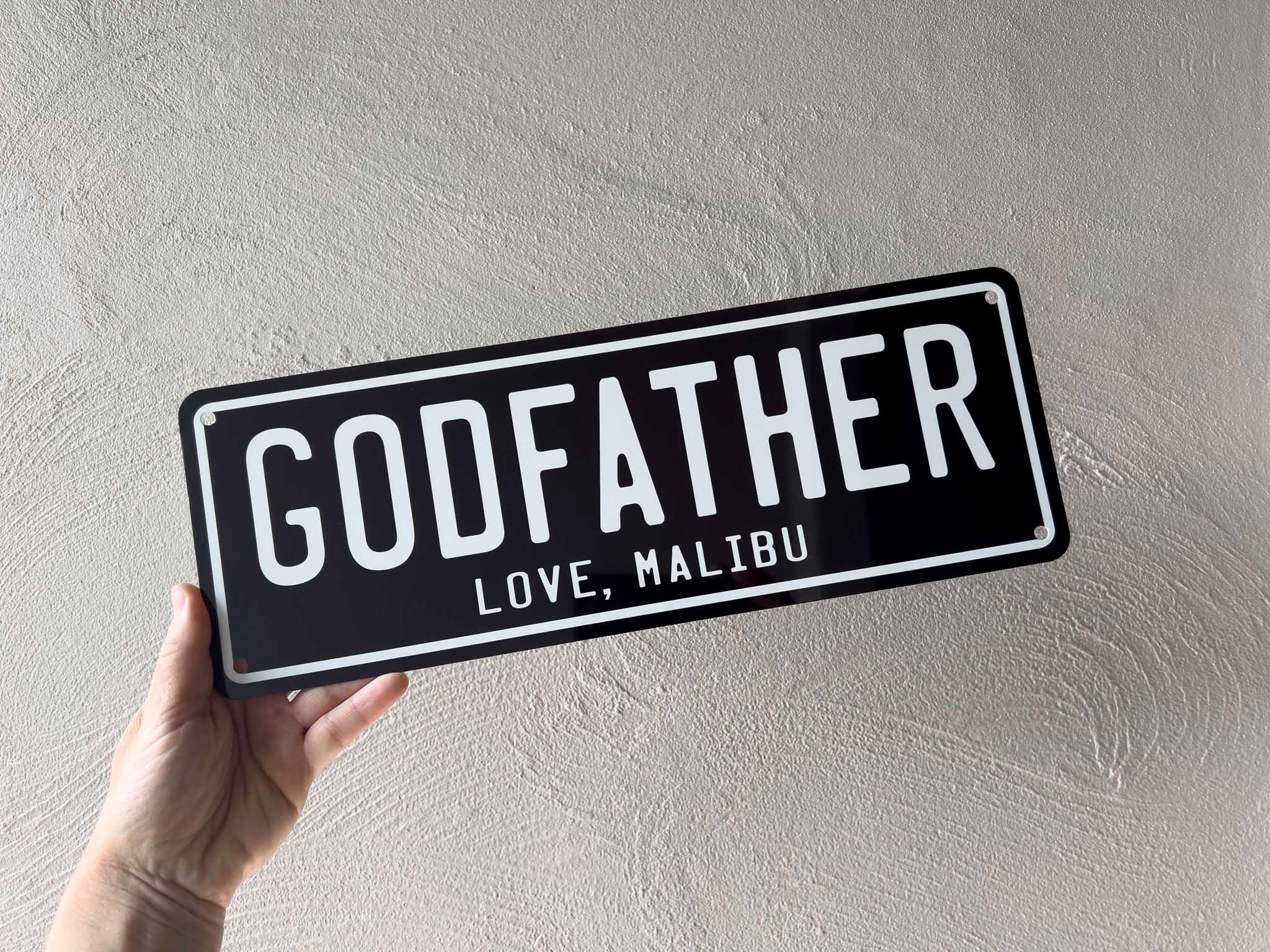 Hand holding a black and white plate with 'GODFATHER' and 'LOVE, MALIBU' text against a light grey background.