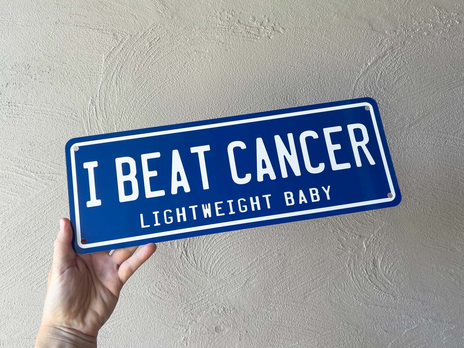 Hand holding a blue sign with 'I Beat Cancer' text against a light gray wall.
