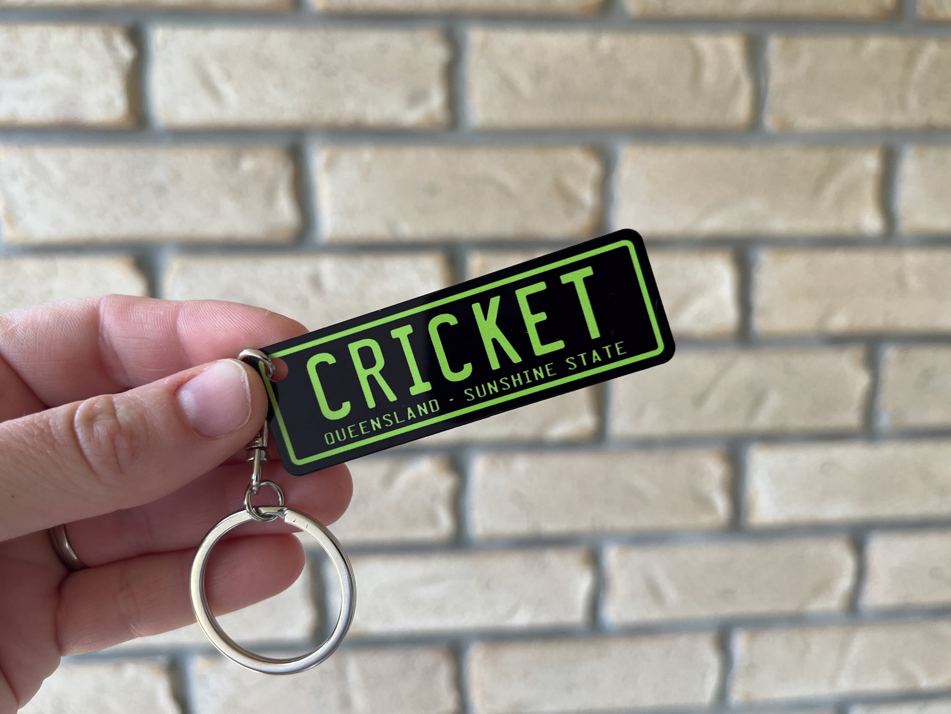 Keychain with 'CRICKET' and 'Queensland' text held against a brick wall.
