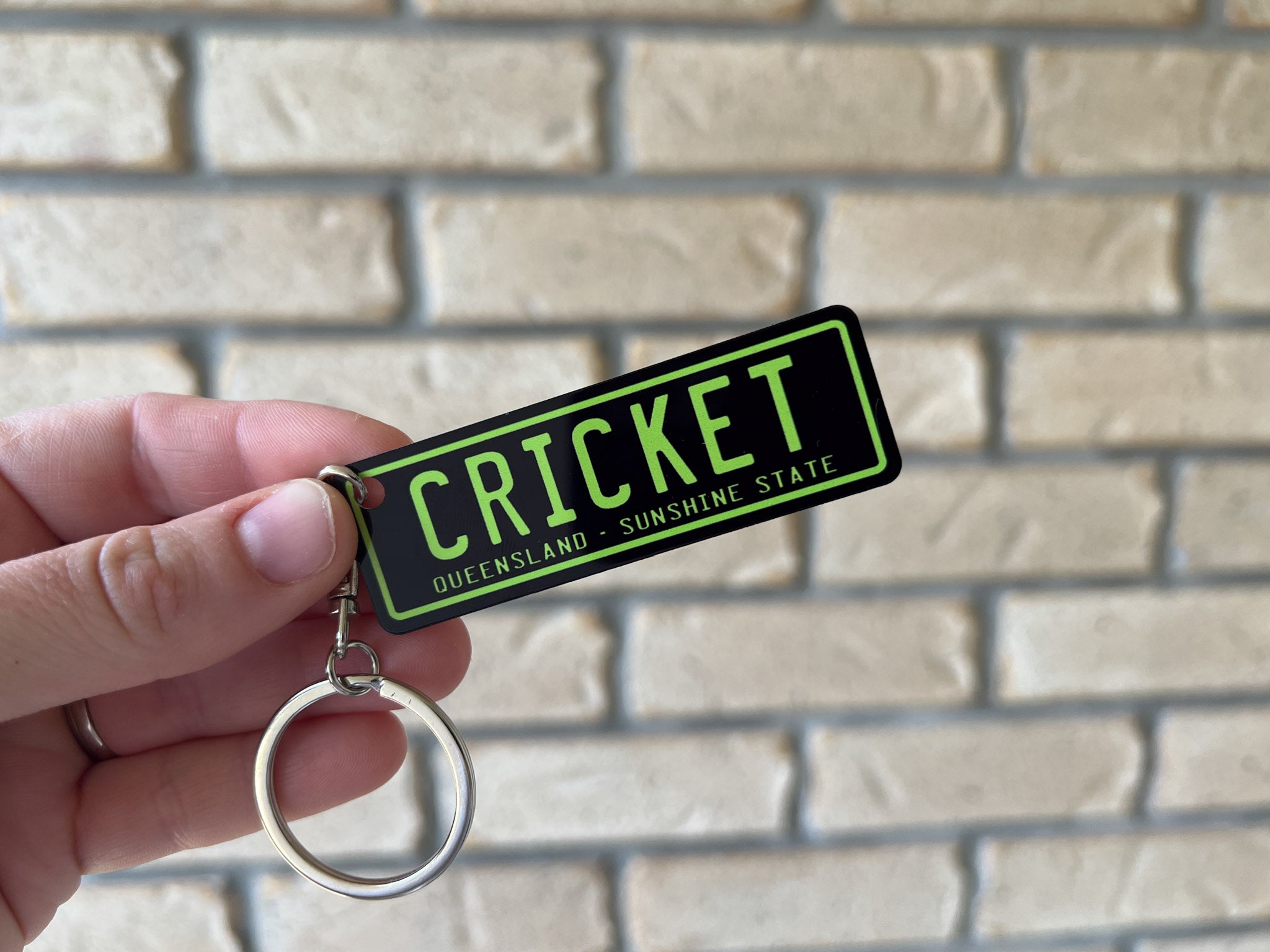 Keychain with 'CRICKET' and 'Queensland' text held against a brick wall.