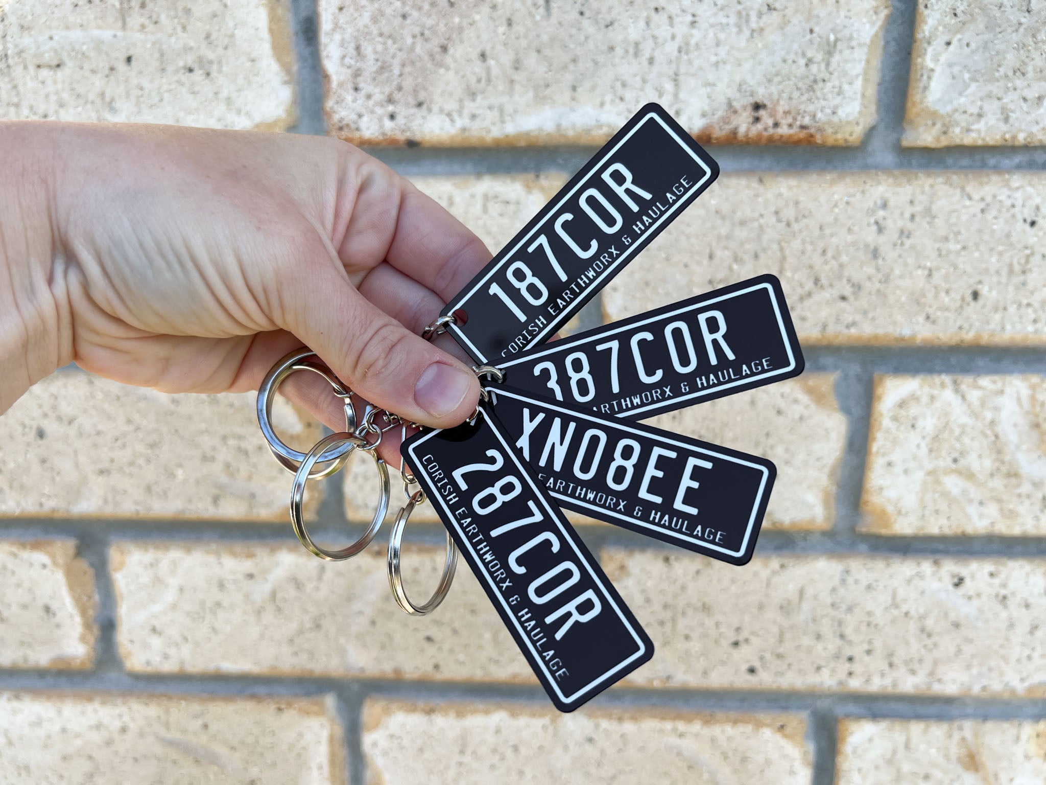 Hand holding four Earthworx & Haulage keychain with a black and white license plate print against a brick wall background