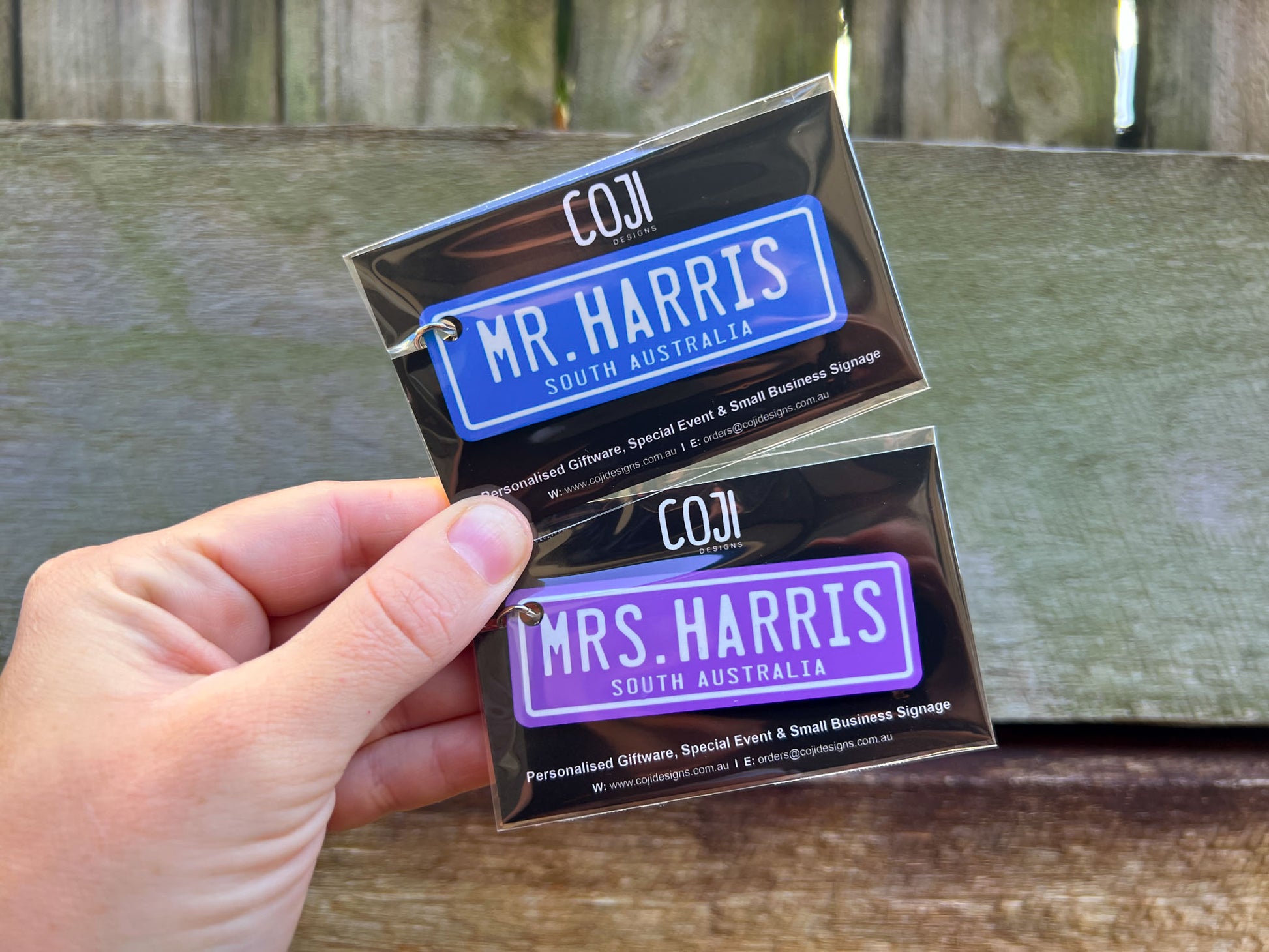 Two metal number plate keychains for 'Mr. Harris' and 'Mrs. Harris' held by a hand against a wooden background.