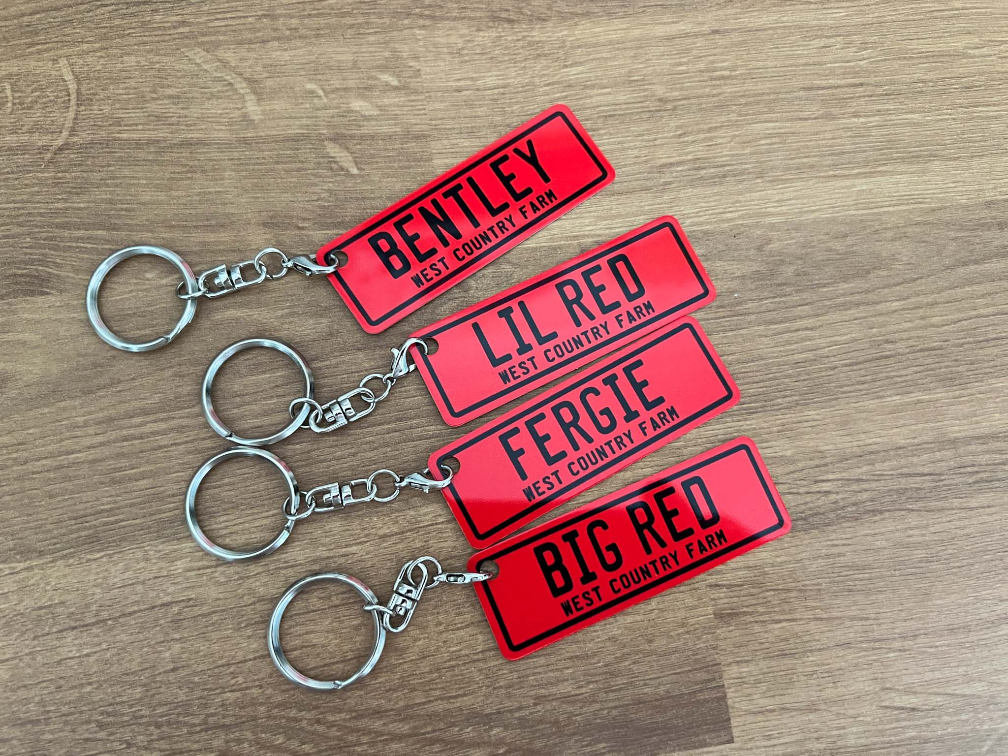 Set of red aluminium keychains for West Country Farm with different names on a wooden surface