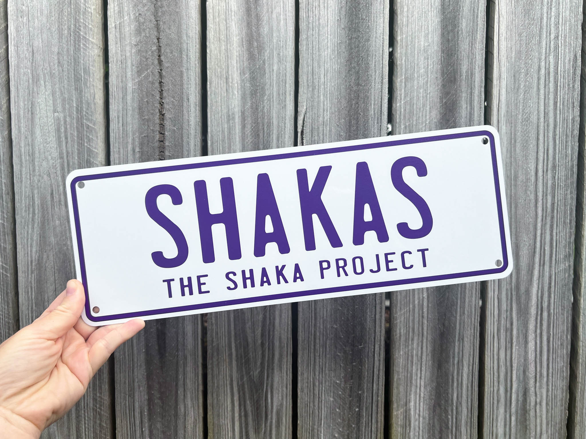 Hand holding a white and purple sign with 'SHAKAS THE SHAKA PROJECT' against a wooden background